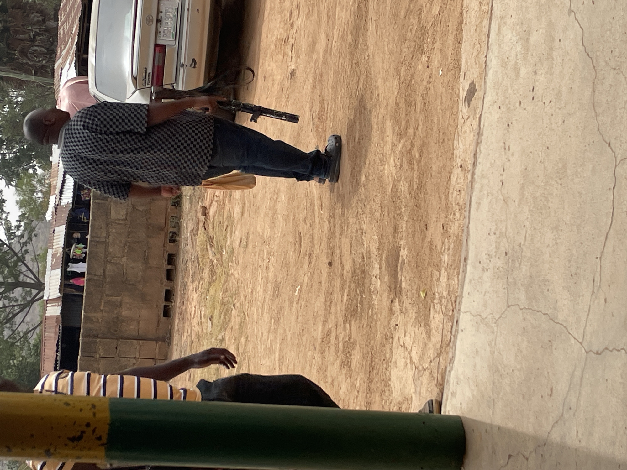The image depicts an outdoor scene, likely in a semi-urban or rural area, consistent with the provided location context of Jos, Nigeria. The time of day appears to be daytime, with diffused light.
The central figure is a man with a bald or closely shaved head, wearing a black and white checkered short-sleeved shirt, dark trousers (likely jeans), and black Crocs-style shoes. He is holding what appears to be a rifle in his right hand, pointing towards the ground, and a light brown bag in his left hand. He stands on sandy, dry earth.
Behind this man, a light-colored (possibly silver or white) Toyota sedan is parked, showing its rear. A license plate is partially visible, bearing the text "PCH 857". The "Toyota" logo is also discernible on the vehicle. Another individual, wearing a light-colored shirt (possibly pink), is partially visible behind the car and the main figure.
In the foreground, a person wearing a light orange/yellow and dark striped shirt is partially visible, with an arm and hand extending into the frame. This individual is largely obscured by a green and yellow barrier or pole that runs across the lower part of the image. The ground in the foreground transitions from sandy earth to a concrete surface with visible cracks.
The background features simple, low-rise structures made of concrete blocks, some topped with corrugated metal roofs. Clothes are hanging to dry on one of these buildings. Sparse trees and vegetation are visible further in the distance. The presence of the rifle is a notable detail.