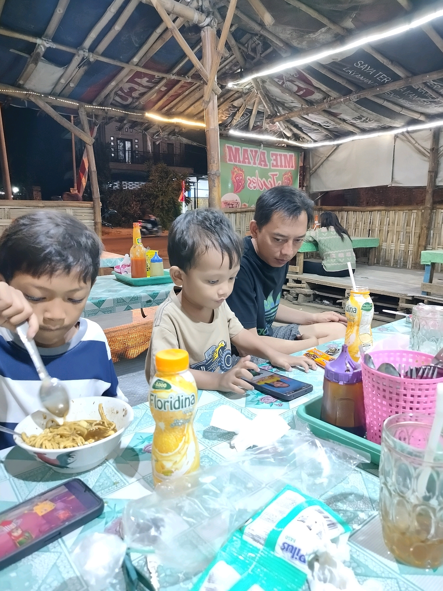 The image captures a dimly lit outdoor dining scene at "Maya Ajim Mie Ayam," a restaurant in Trowulan, Indonesia, as indicated by the signage. Two young boys and a man are seated at a table covered with a patterned tablecloth. The boy on the left is actively eating noodles from a bowl with a spoon, while the boy on the right is looking at a smartphone. The man is also looking down, possibly at his own phone or the food. The table is cluttered with various items including a bottle of "Floridina" drink, a glass with ice, a pink basket holding utensils, and plastic bags. In the background, a woman is visible sitting at another table, and the restaurant's rustic bamboo structure and thatched roof are prominent features of the environment. The overall atmosphere suggests a casual, family-friendly dining experience.