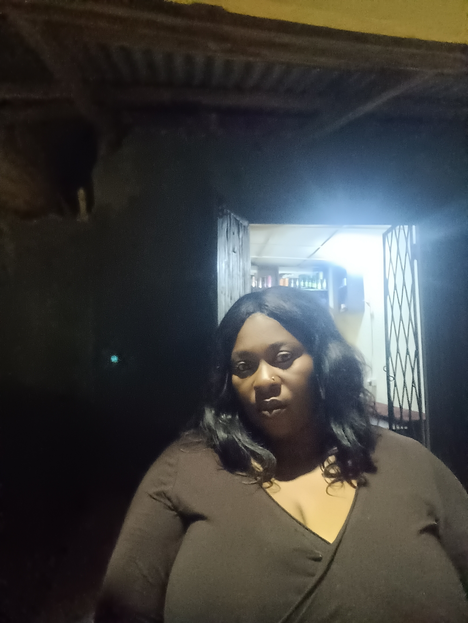 A woman stands in the foreground in a dimly lit environment. She is wearing a brown v-neck garment. Behind her, a door with a metal security gate is open, revealing a brightly lit interior with shelves stocked with various items. The location is possibly a shop or store. Over the entrance way is a dark overhang. While the request asks for the location in the city of Jos, Nigeria, this detail is not explicitly confirmed within the image.