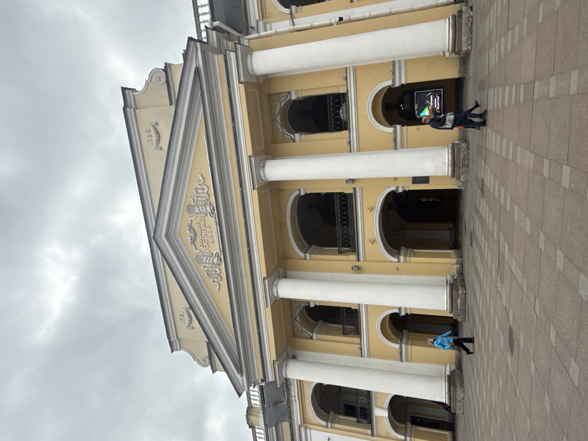 The image, captured in a portrait orientation but showing a landscape scene rotated 90 degrees clockwise, features a grand, light yellow neoclassical building under an overcast sky. The building is characterized by a prominent portico with white columns supporting a pediment. On the pediment, decorative relief work includes a ship motif and the Russian text "БОЛЬШОЙ ГОСТИНЫЙ ДВОР" (Bolshoy Gostiny Dvor), with the year "1785" ornately displayed below it.
The building's facade is punctuated by numerous arched entrances and windows, some of which are recessed and feature dark, decorative railings. The ground in front of the building is a paved square, with a pattern of light brown and off-white tiles.
Two individuals are visible on the paved area. On the right, a person in a dark jacket and trousers stands near an arched entrance, appearing to look at a large digital screen. The screen displays colorful graphics and partially legible text, including "ГОСТИНЫЙ ДВОР" and "НОКТЮРН". Further left, another person, dressed in a bright blue jacket and dark trousers, walks across the paved area.
The sky is a uniform, pale grey, indicating an overcast day. The architectural style, the visible text in Russian, and the historical date confirm the location as the Gostiny Dvor in Saint Petersburg, Russia. The scene depicts a public square or street in front of this notable landmark.