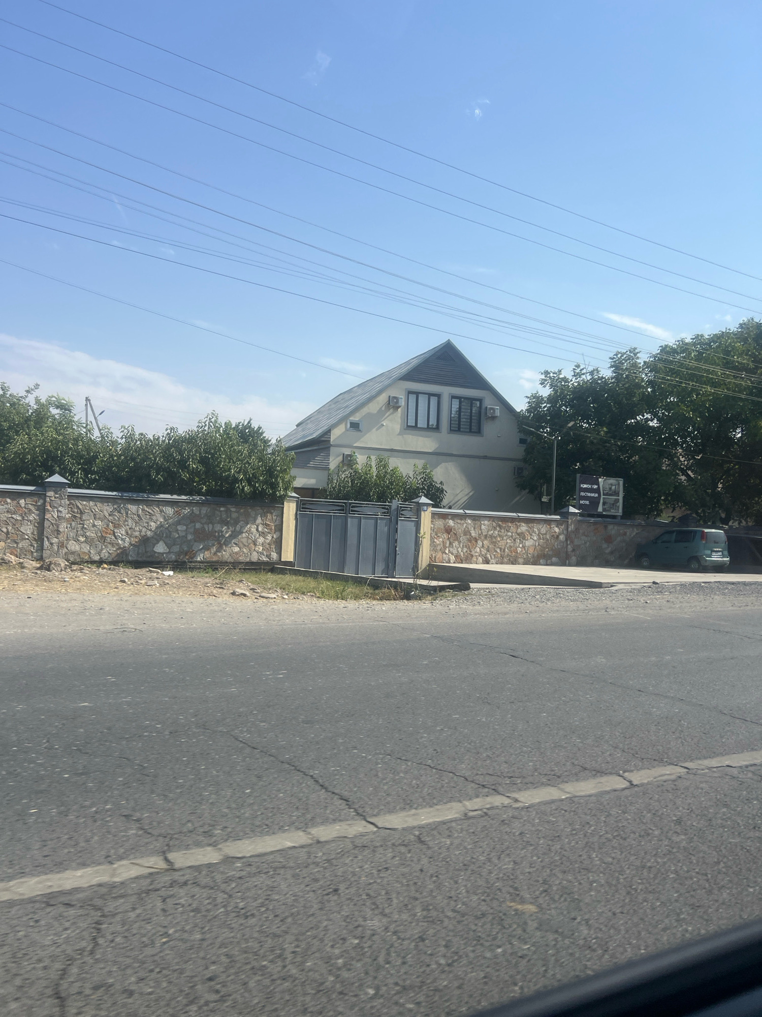 The image shows a street scene in Özgön, Kyrgyzstan, on a clear, sunny day. In the foreground, a cracked asphalt road runs horizontally across the frame. Behind the road, a stone wall topped with a grey border encloses a property. A grey metal gate is visible in the wall, with a concrete ramp leading up to it. A light-colored building with a steeply pitched roof and dark trim is situated behind the wall, featuring two windows and air conditioning units. Lush green trees and bushes surround the property. A small teal-colored car is parked on the right side of the property, near a sign that reads "KONON YUR GOSOTINBER HOTEL". Power lines are strung across the bright blue sky above.