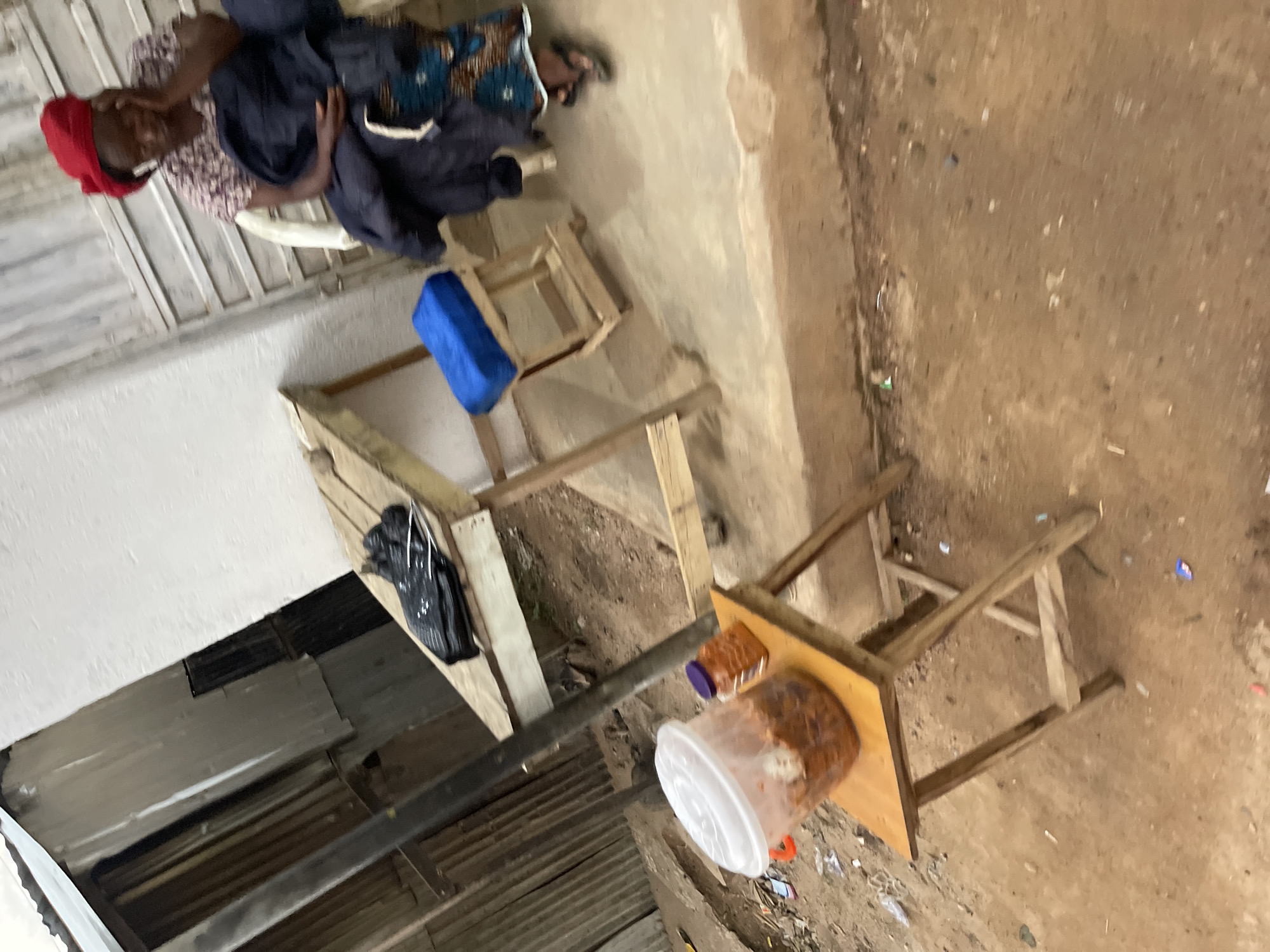 The image depicts an outdoor scene, possibly a market stall, in Jos, Nigeria. A person is partially visible, wearing a red head covering and a patterned shirt, and appears to be seated. Next to them, there is dark blue fabric and a blue object, possibly a cushion or fabric. In the foreground, a wooden stool or table holds two containers: a small one with a dark lid and a larger, clear plastic container with a white lid, filled with what appears to be food items. The ground is rough and sandy, with some debris scattered around. To the left, a white wall and corrugated metal sheeting suggest a makeshift structure. The lighting indicates it's daytime, and the overall impression is of a functional, perhaps informal, vending area. The image is oriented sideways.