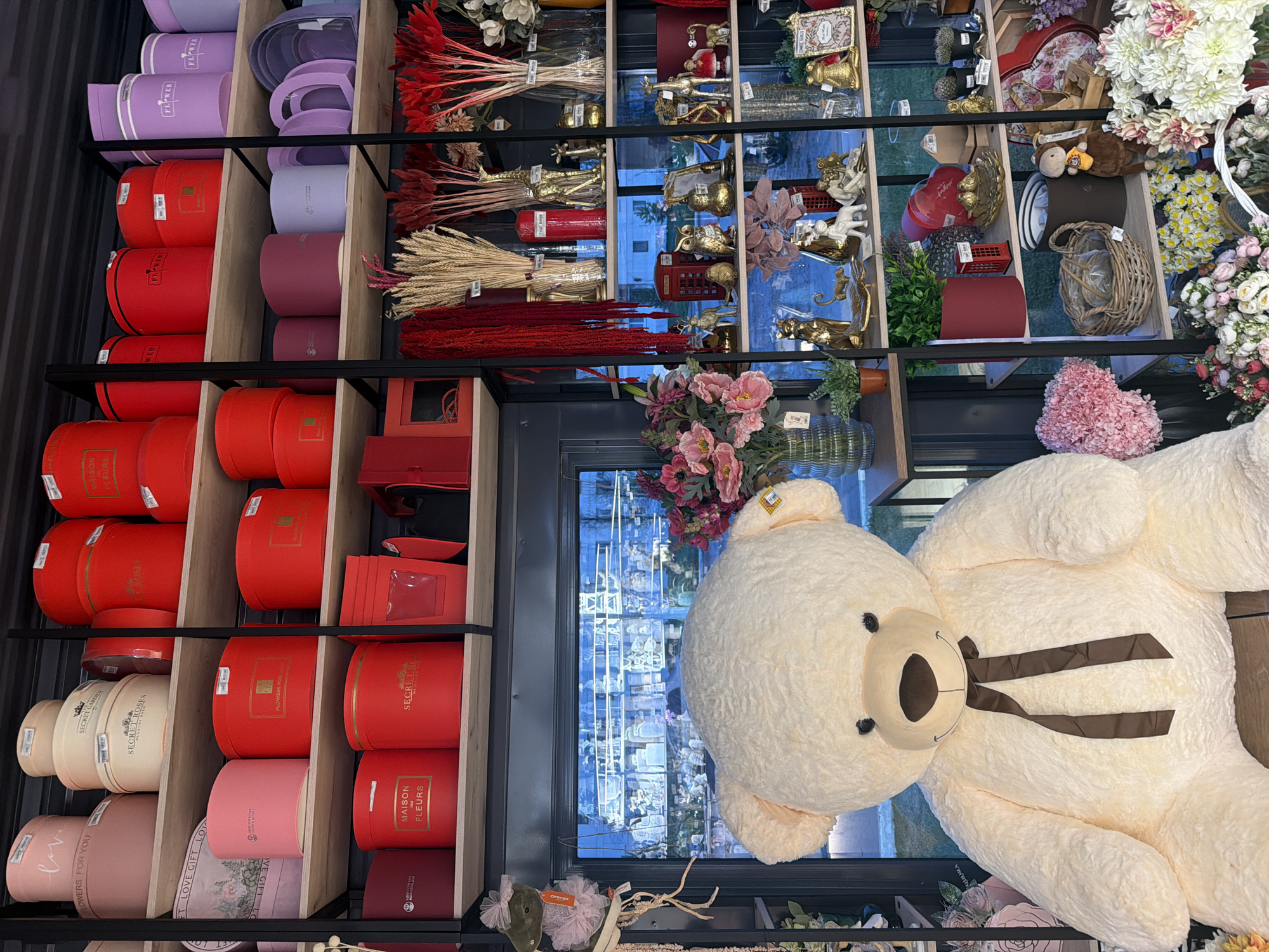 Here is a detailed description of the image:
**General Overview:**
The image appears to be taken inside a store, likely a flower shop or a gift shop that also sells flowers. The photograph is taken from an elevated perspective. The composition includes shelving units stocked with various items, and a large teddy bear.
**Location and Time:**
The location is Chaikovsky, Russia. The time is daytime.
**Subjects and Objects:**
* **Shelving Units:** There are wooden shelving units filled with various items. On the left side, there are stacked round boxes in red, pink, and beige, possibly for flower arrangements or gifts. On the right side, the shelves contain dried flowers, vases, decorative figurines (including golden reindeer and people), and other decorative objects.
* **Teddy Bear:** A large, plush teddy bear is positioned near a window. It has a light-colored fur, a brown bow tie, and black eyes.
* **Flowers:** There are fresh flowers, including pink roses.
* **Window View:** The reflection in the window provides a view of the street and surrounding buildings. The buildings appear to be modern.
**Environment:**
* **Lighting:** The environment is well-lit, with both natural light coming from the window and likely artificial lighting within the store.
* **Atmosphere:** The setting appears to be a retail environment with a focus on gift items and floral arrangements.