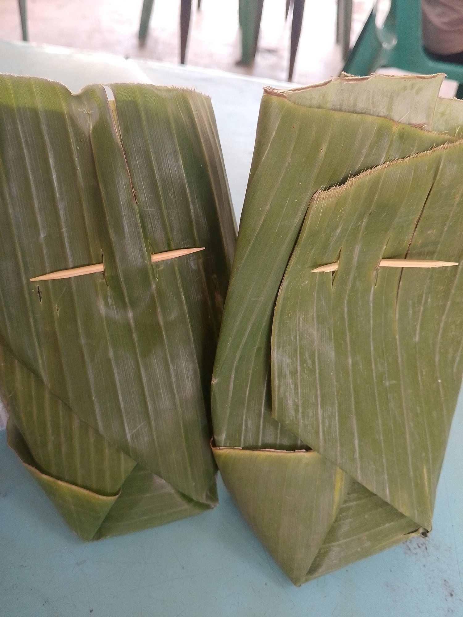 Two portions of food are wrapped in green banana leaves. A small wooden toothpick holds each leaf in place. The items sit on a light-blue surface. In the background, hints of chairs can be seen. The location is inferred to be in Lhokseumawe, Indonesia.
