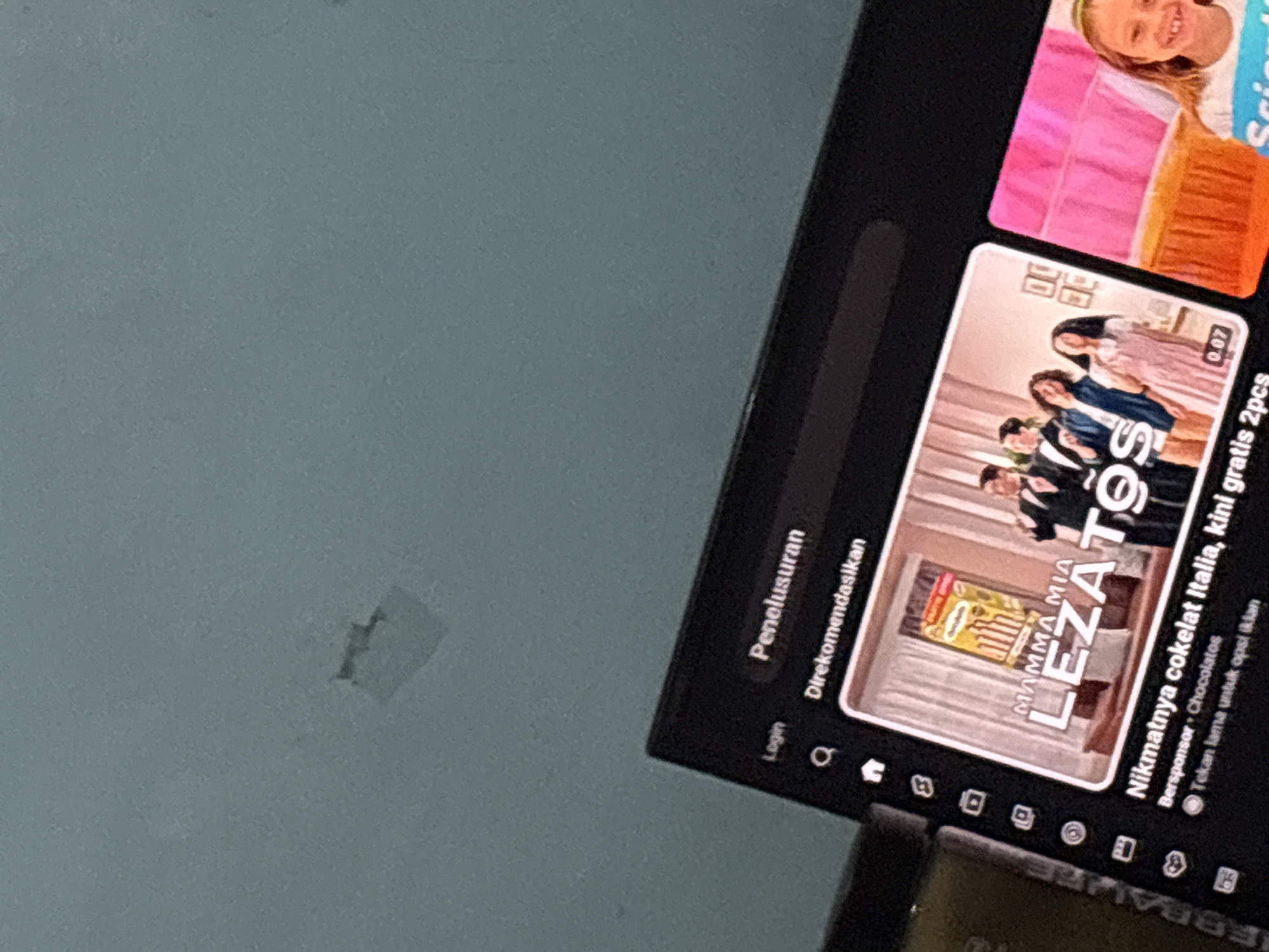 The image shows a close-up of a tablet screen displaying a streaming service interface. The screen is angled upwards towards a plain, light-colored ceiling. On the left side of the tablet screen, a vertical menu bar with icons is visible. The main content area shows two video thumbnails. The left thumbnail features a group of people in formal wear posing in front of a product box labeled "MAMMA MIA LEZATOS" and text that appears to be an advertisement for Italian chocolate. The right thumbnail shows a young girl smiling in front of a bright pink background. The lighting in the scene is dim, and the image quality suggests it was taken with a mobile phone. No specific location in Danawinangun, Indonesia can be determined from the visual information provided.