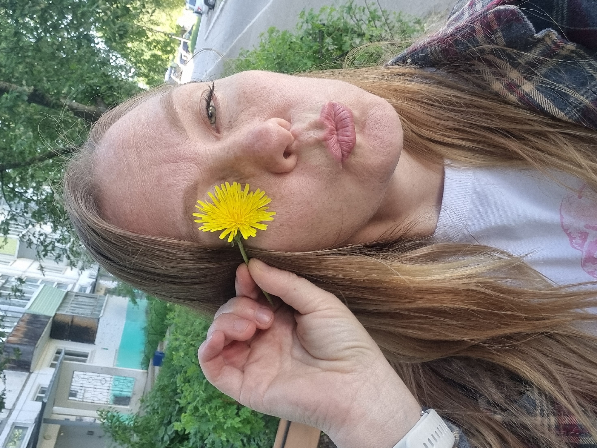 A light-skinned woman is lying down, holding a yellow dandelion flower next to her face. She is wearing a white T-shirt and a plaid shirt, and has a white smartwatch on her wrist. The background includes trees, greenery, a multi-story building, and a street. Based on the architecture and vegetation, the location is Chaikovsky, Russia.