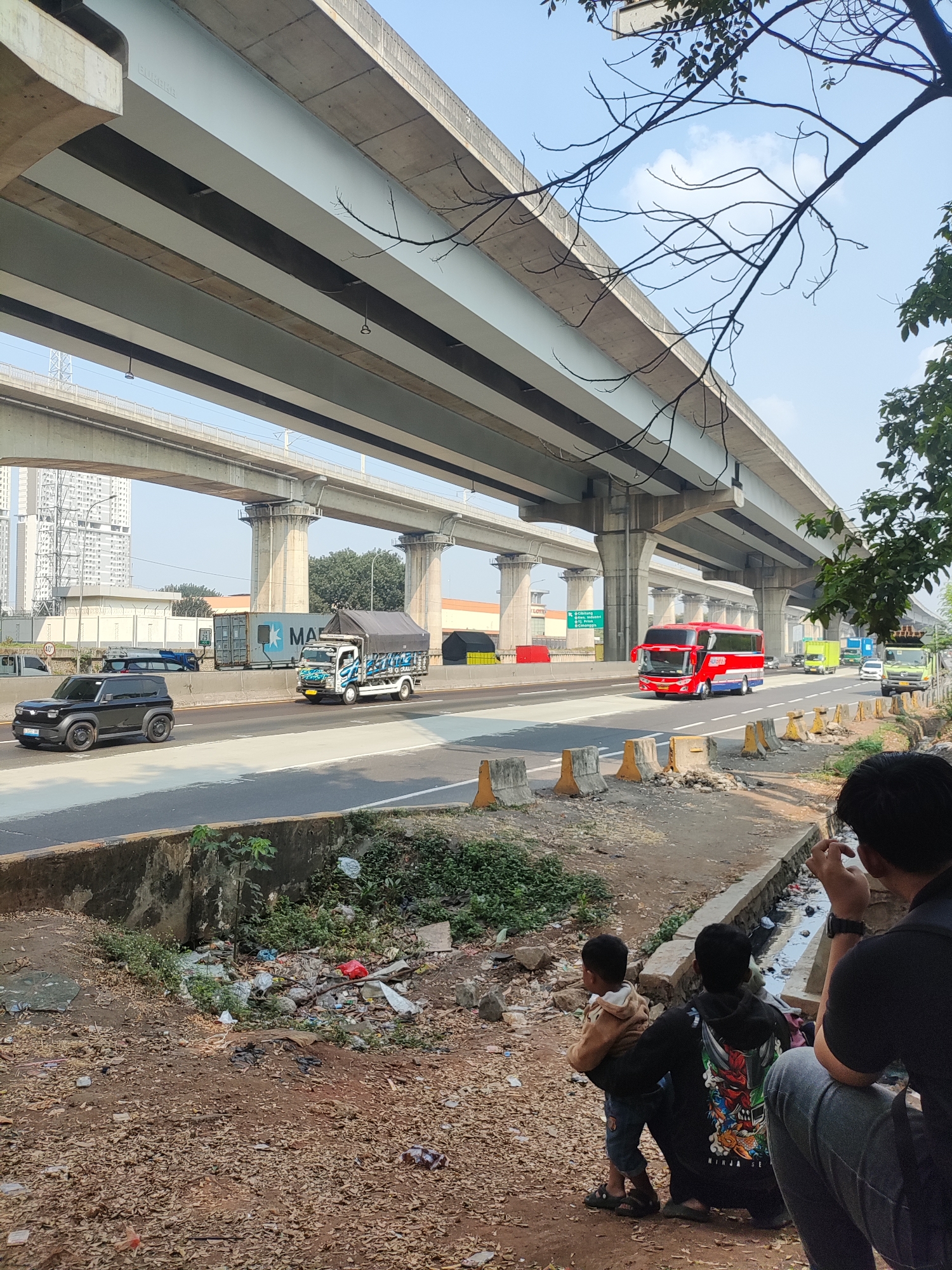 In Gandamekar, Indonesia, under a multi-level highway overpass, a roadside scene unfolds. A black SUV drives on the elevated highway, followed by a white truck covered with a dark tarp, and a red double-decker bus. The highway is lined with concrete barriers and traffic cones. In the foreground, two children and a man are seated on the ground. The ground is covered with fallen leaves and debris, with a narrow ditch running alongside a concrete embankment that is filled with trash and overgrown vegetation. The scene captures a slice of daily life under the sprawling infrastructure of Gandamekar.