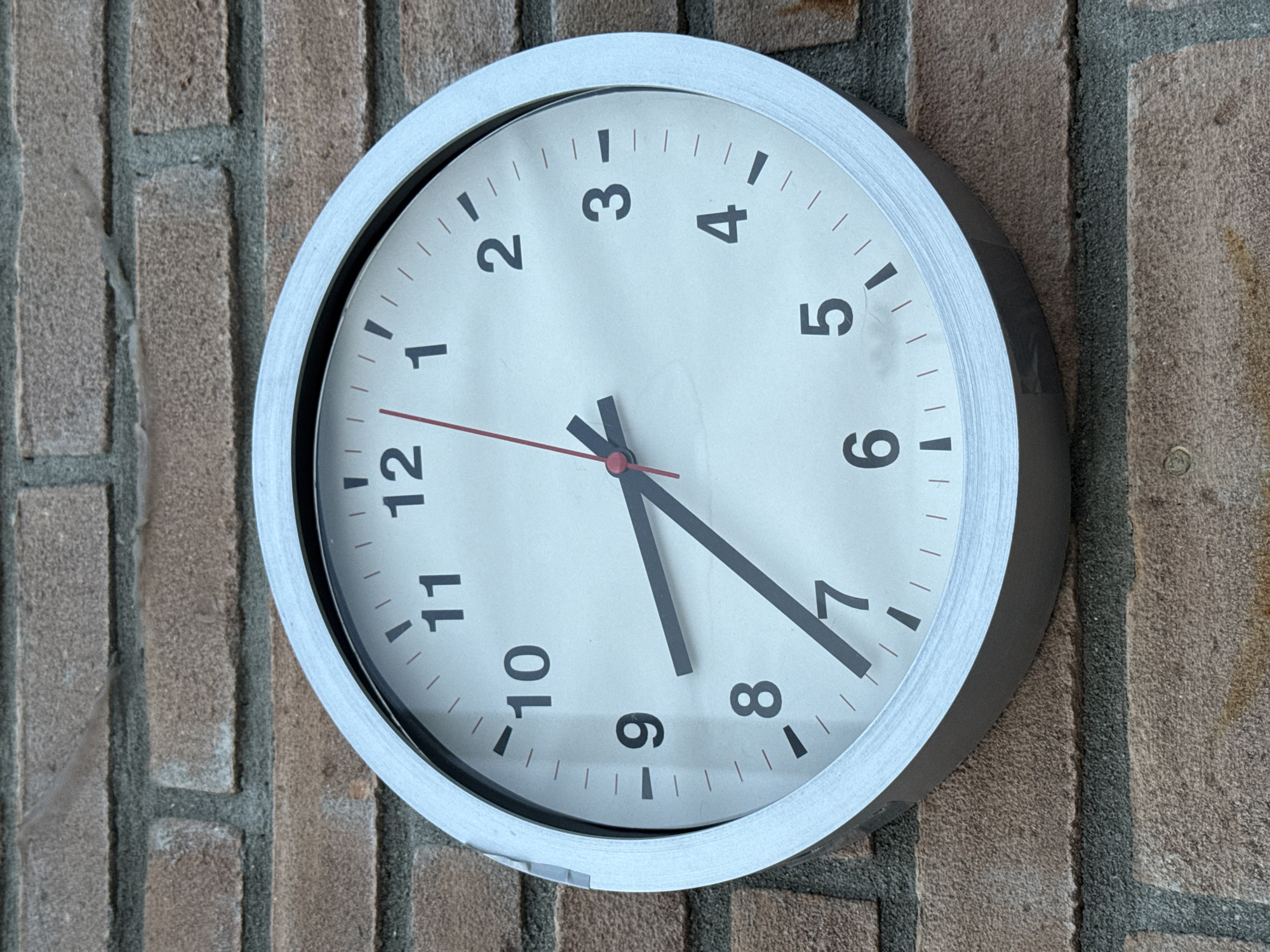 A round analog clock with a silver frame is mounted on a brick wall. The clock face is white with black numbers and hour markers. The hour hand is pointing between 8 and 9, closer to 8. The minute hand is pointing towards 7. The second hand is red and points to the 12. The brick wall is made of reddish-brown bricks with gray mortar. The scene does not indicate any specific location in the city of Parma, Italy.