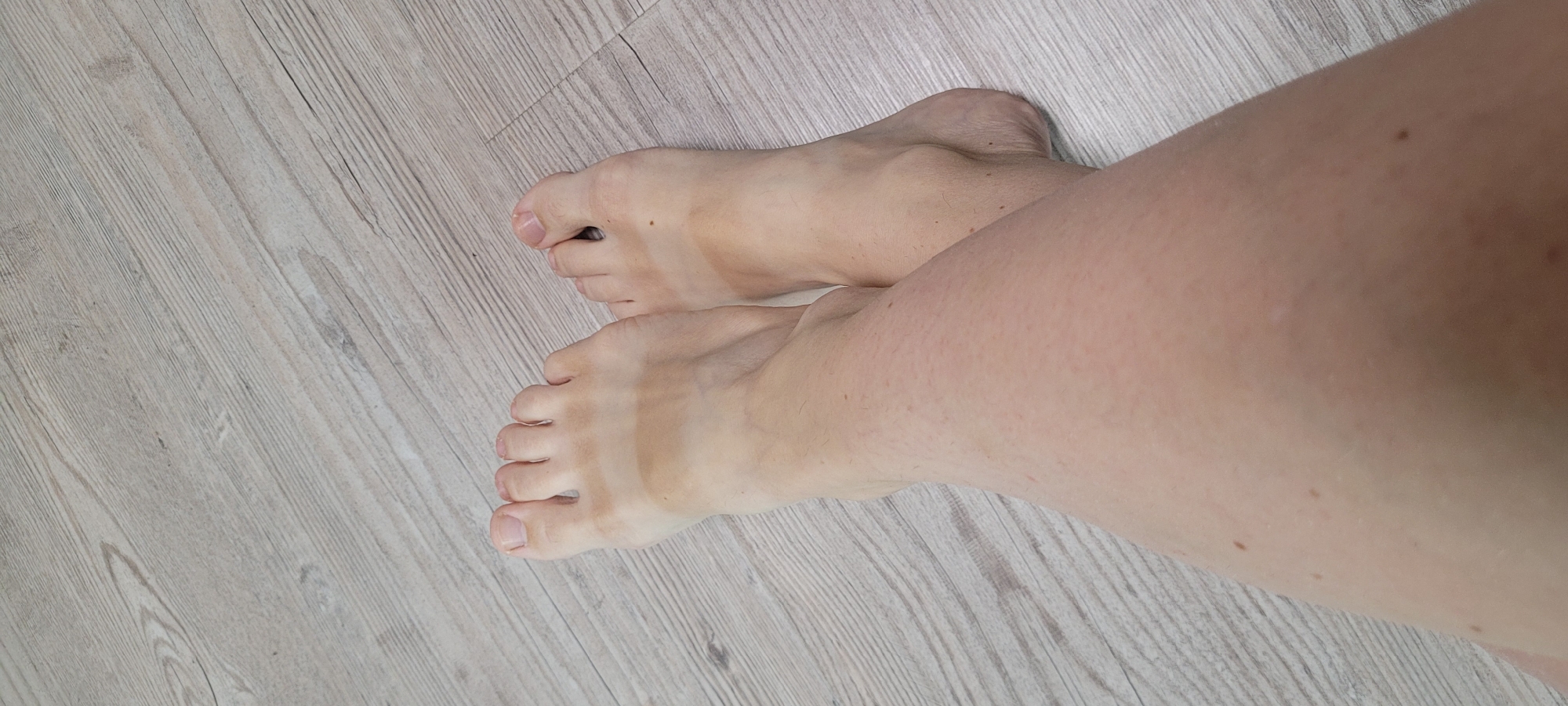 The image shows a close-up of a person's legs and feet resting on a light-colored wooden floor. The feet are bare, and there are noticeable tan lines across the tops of both feet, indicating they were exposed to the sun while the rest of the feet were covered. The skin appears fair with some freckles and minor blemishes. The wooden floor has a distressed texture with visible grain and lines. The lighting is even, suggesting an indoor environment. No specific location details are discernible from the image itself.