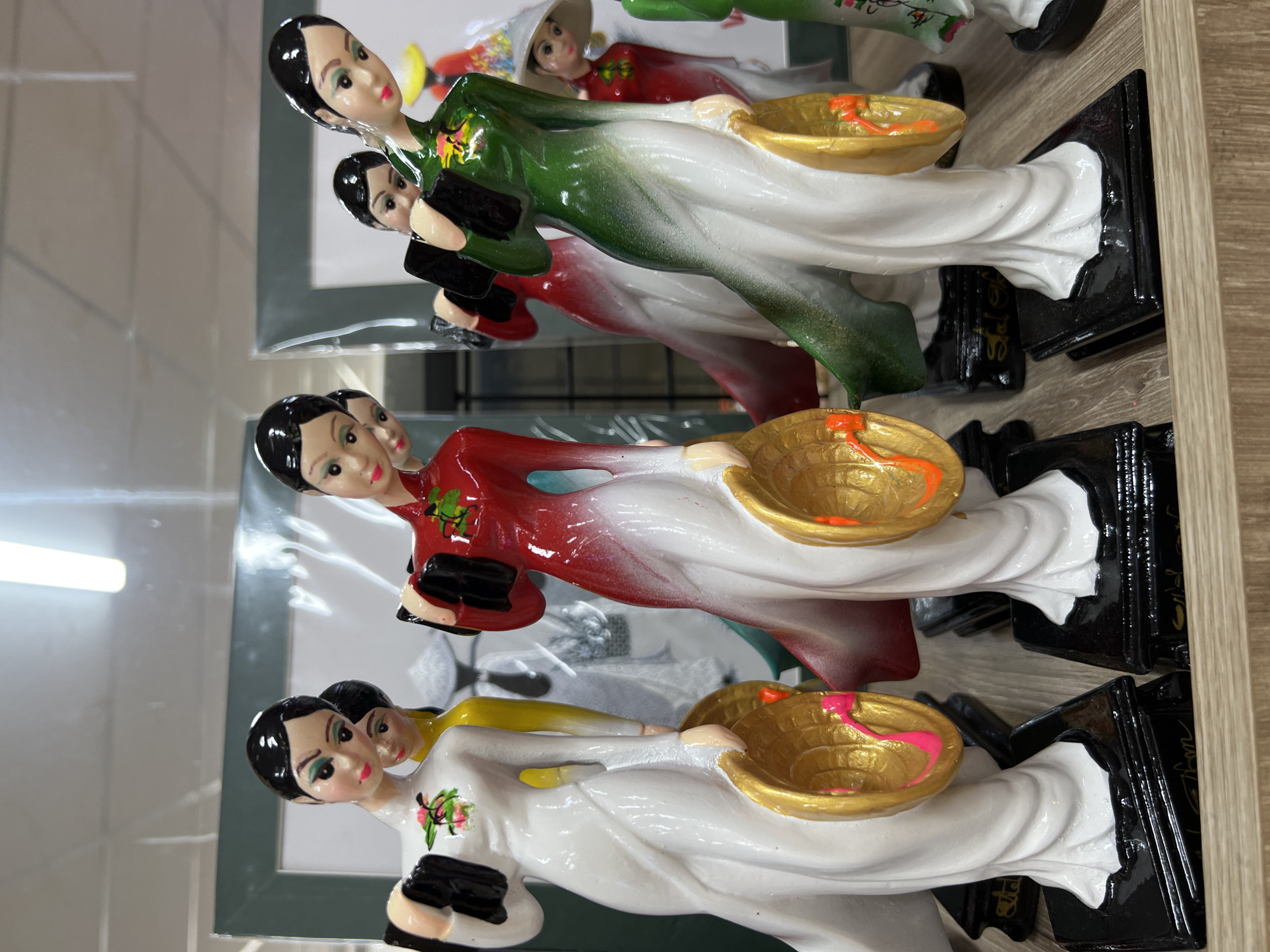 The image shows a close-up, eye-level shot of several decorative figurines displayed on a wooden shelf. The figurines are of women in traditional Vietnamese attire, likely Áo dài, holding conical hats and what appear to be small books or wallets. The figures are made of a glossy material, possibly ceramic or resin, and are painted in vibrant colors. The primary figures in the foreground are dressed in white, red, and green dresses, with elaborate detailing. The backgrounds suggest a retail environment, with framed pictures and other merchandise visible, though the focus remains on the figurines. The lighting appears to be from overhead fluorescent fixtures, casting a slight glow on the polished surfaces of the figures. The overall impression is that of a display for sale in a shop. The scene is likely located in Nha Trang, Vietnam, given the cultural context of the figurines.