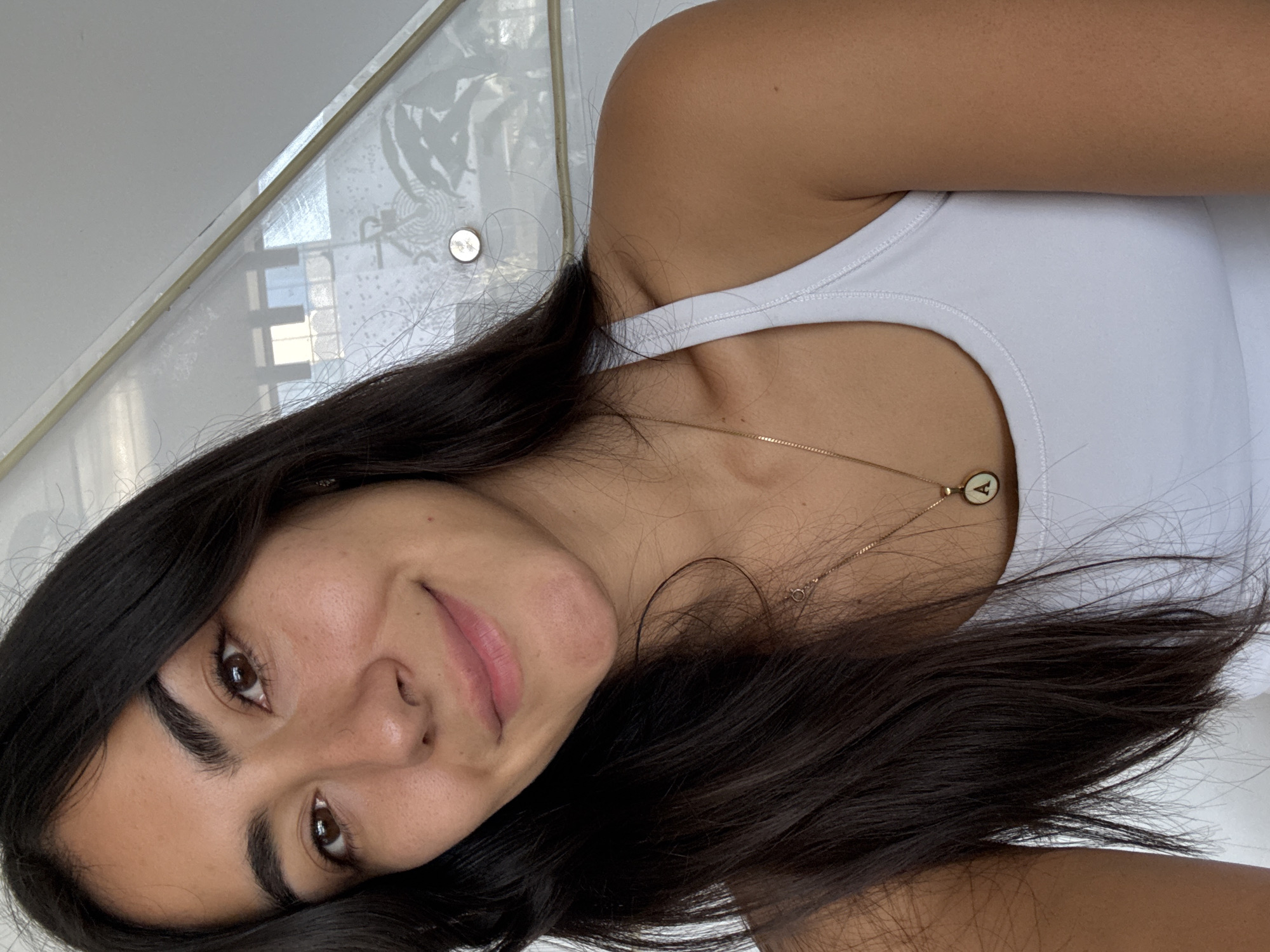 This bright, naturally lit portrait captures a young woman from Puebla, Mexico, smiling gently towards the camera. She has long, dark brown hair that frames her face and flows over her shoulders, and expressive dark brown eyes. Her complexion is warm, and she wears a subtle, natural expression.
She is dressed in a plain white tank top. A delicate gold chain necklace is visible around her neck, featuring a small, circular pendant with a white inlay and a clear black letter "A" inscribed on it.
The background is bright and predominantly white, possibly a wall or ceiling. Behind her head and left shoulder, there is a reflective surface, possibly a window or glass, showing blurred reflections of what appear to be window frames or structural elements, along with some indistinct patterns. A small, round, metallic object is also visible in this reflected area. The image is captured with the subject appearing to be lying down or the camera angled steeply, creating an intimate perspective. The lighting suggests it is daytime.