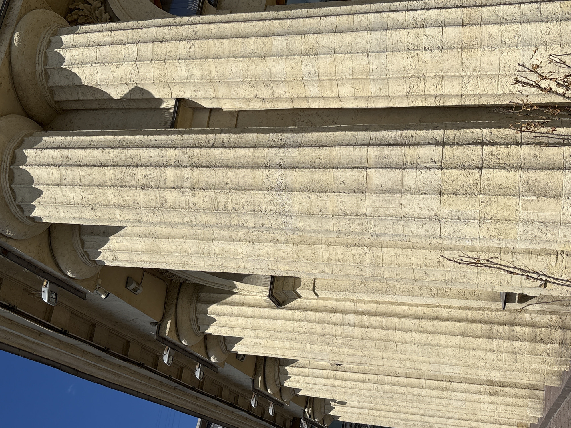 The image shows a close-up, tilted view of fluted columns and architectural details of a building, likely in Saint Petersburg, Russia. The columns are made of a light-colored stone, possibly sandstone or limestone, and are heavily weathered, showing signs of age and exposure to the elements. The fluting is deep and well-defined. In the upper portion of the image, sunlight casts strong shadows on the columns, indicating it is daytime, likely with clear weather. A dry, leafless branch from a tree is visible extending across the top right of the frame, adding a natural element to the man-made structure. Below the columns, the underside of an overhang is visible, featuring a series of modern, white light fixtures and what appear to be security cameras or sensors attached to a dark metal railing. The clear blue sky is visible at the bottom left of the image. The overall impression is one of grand, classical architecture contrasted with utilitarian modern additions. There is no discernible text in the image.