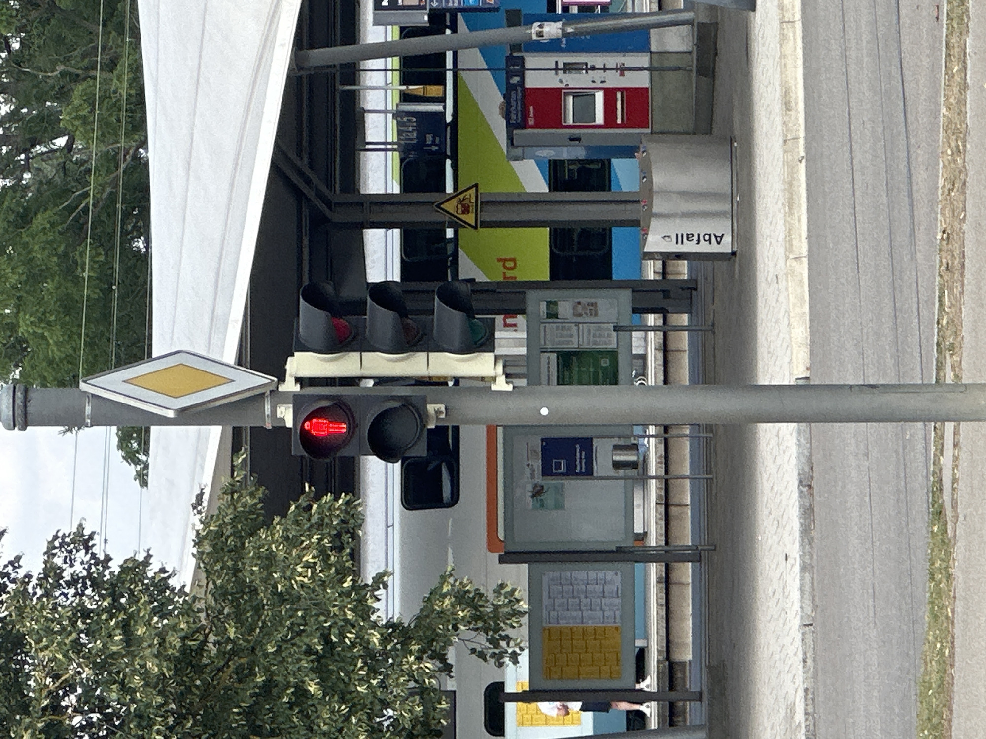 The image shows a red pedestrian traffic signal indicating "don't walk" in front of a train station platform in Günzburg, Germany. A modern train with blue and green accents is stopped at the platform. To the left, a person in shorts and a t-shirt stands near a bulletin board. To the right, a ticket machine and a trash receptacle labeled "Abfall" are visible. The scene is overcast, with trees visible behind the station's awning.