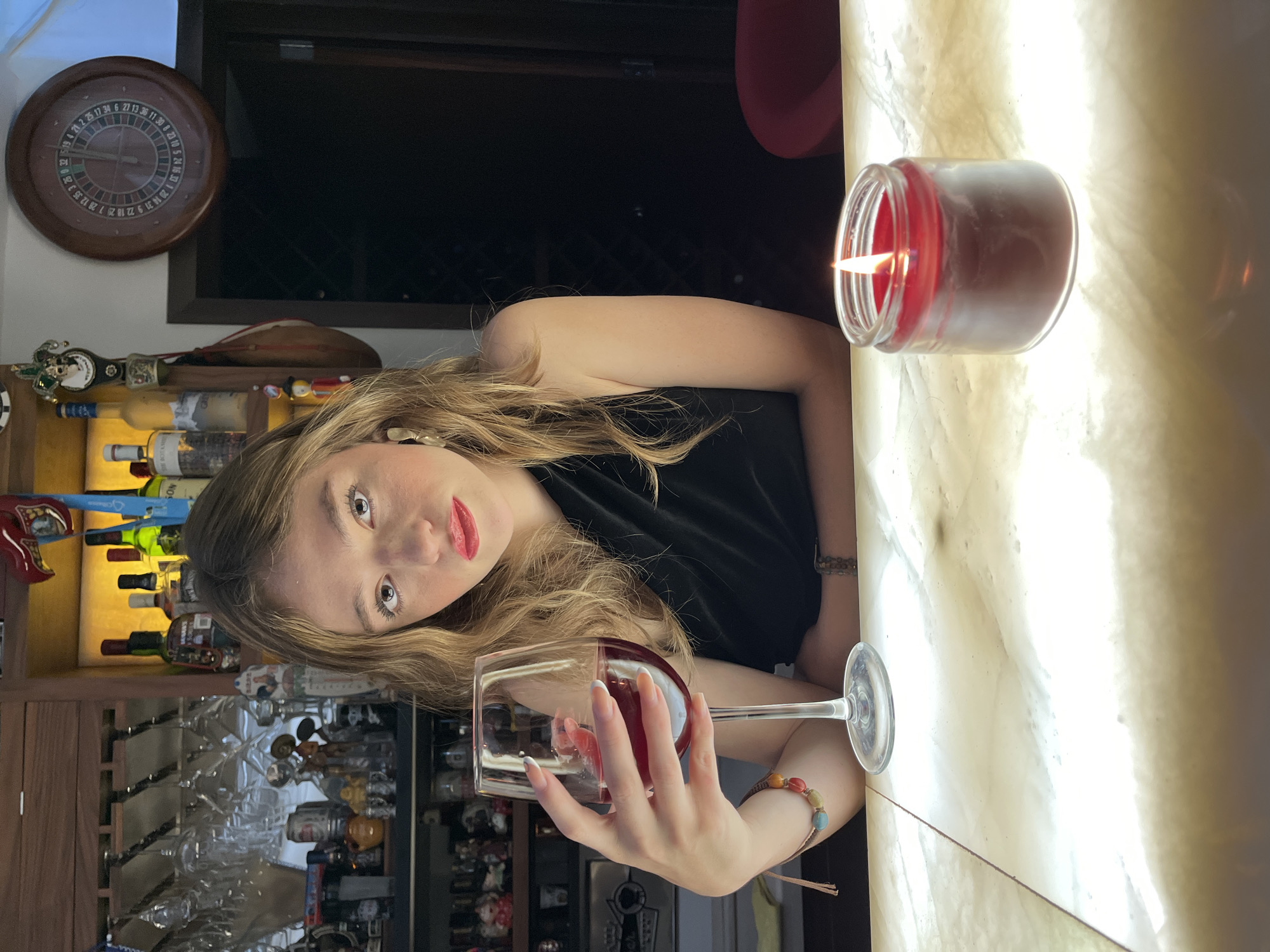 A woman sits at a glowing bar, holding a glass of red wine, and looks directly at the camera. She has long, wavy blonde hair, is wearing a black top, and has red lipstick and long nails with white tips. Bracelets, including one with colorful beads, adorn her wrist. To her right, a red candle in a clear jar burns, casting a warm glow on the translucent bar surface.
The background reveals a well-stocked bar area. Shelves behind the woman hold various bottles, some illuminated from beneath, along with hanging wine glasses and decorative items. On the upper left wall, a wooden roulette wheel-shaped clock is visible. Above the shelves, a wooden paddle or oar is placed. The overall lighting is dim, suggesting an evening or night setting.
Visible text from the roulette wheel includes numbers such as "21 2 25 17 34 6 27 13 36 11 30 8 27" and "10 10 5 24 16 33 1 26 14 31 9 22 18 25 7 28 12 35 3". The partial word "SON" and number "51942" are also discernable, likely from a bottle label. The scene takes place in San Bernardino Tlaxcalancingo, Mexico.
