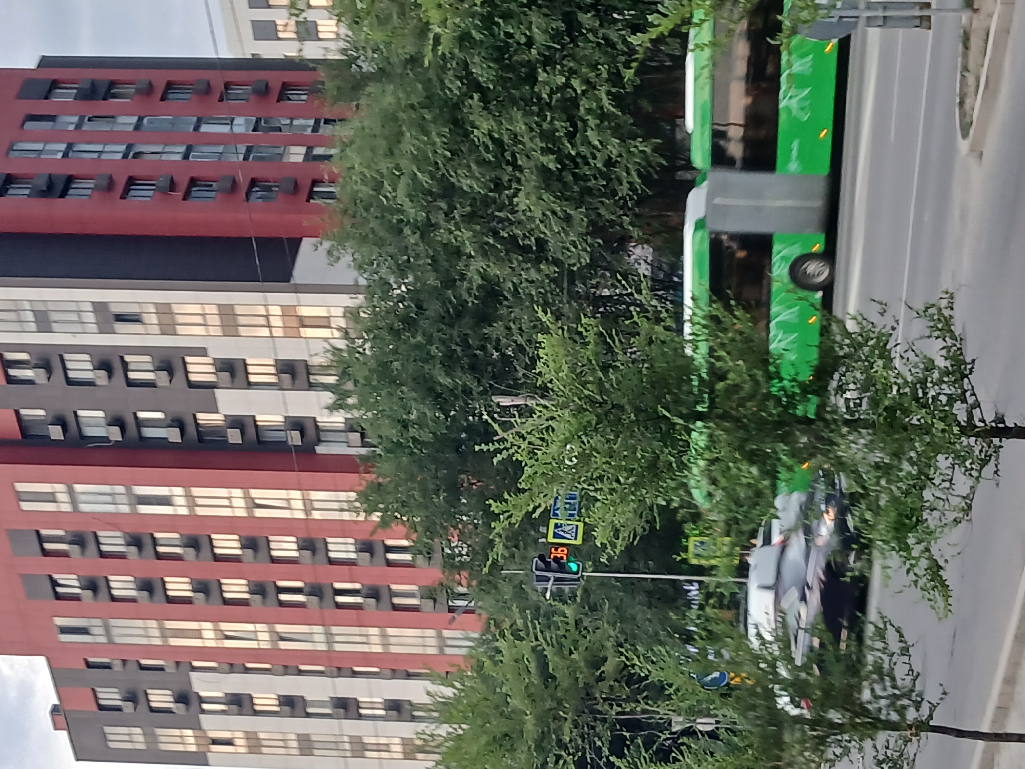 The image depicts a street scene with modern buildings and a bus. The buildings are tall and feature a repeating pattern of windows and balconies. One building is predominantly red with dark gray accents, while another appears to be lighter in color, possibly white or beige, with numerous windows reflecting the sky. The bus is green and white, with a stylized design on its side. It is parked on a paved road.
Several trees with dense green foliage are scattered throughout the scene, partially obscuring the view of the buildings and the bus. In the foreground, there are more trees and branches. A traffic light with a green signal is visible, indicating that traffic is allowed to proceed. There are also road signs, including one with a pedestrian crossing symbol. The overall impression is that of a city environment during daylight hours, possibly late afternoon due to the lighting. The weather appears to be clear. The image is taken from a slightly elevated perspective, looking down on the street. There are no discernible people in the image.