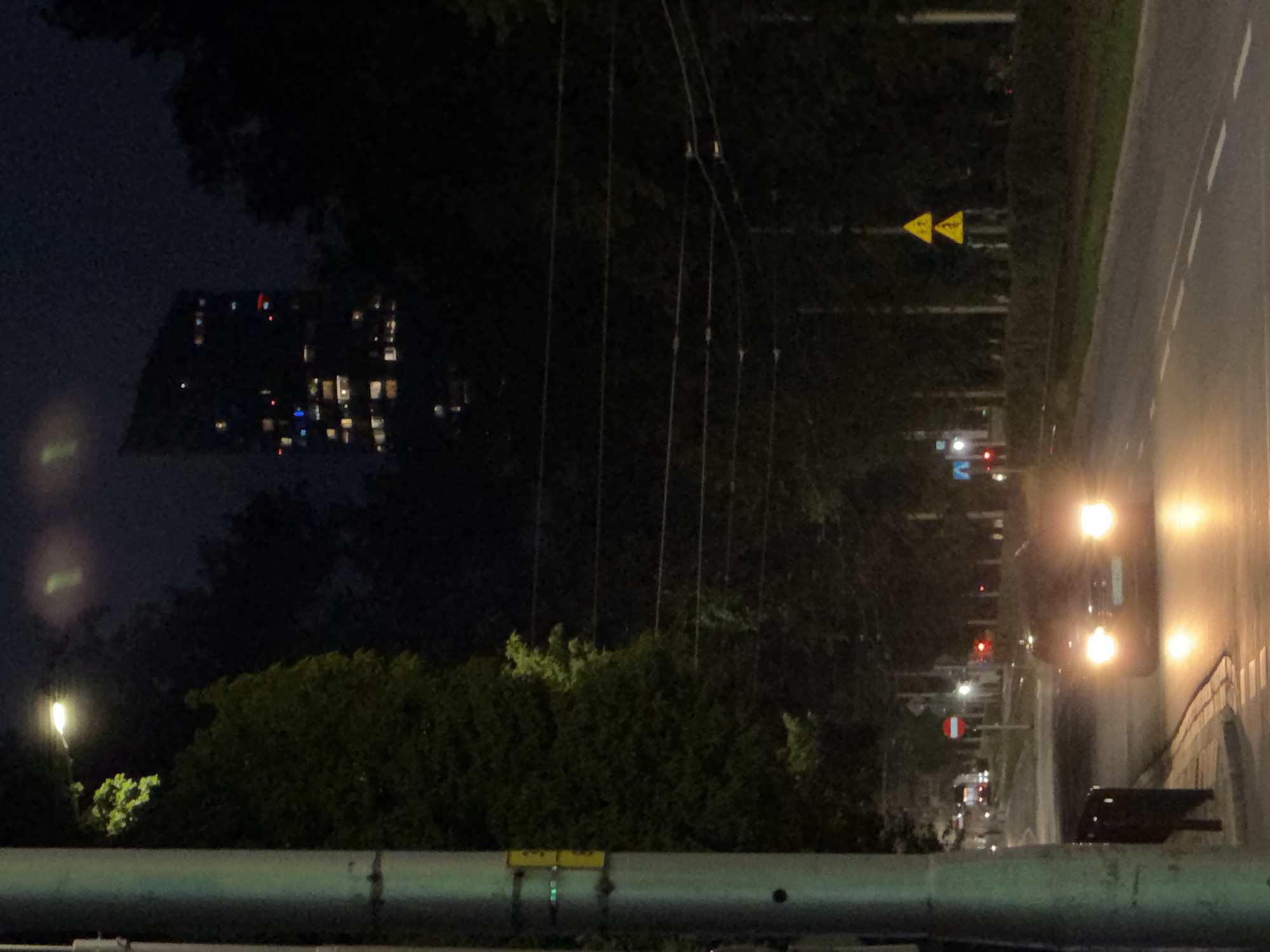 The image depicts a nighttime street scene in an urban environment. In the foreground, a street lamp stands on the left, and a dark-colored car with its headlights illuminated is driving on the road. The road is paved and has lane markings. On the right side of the road, there are two triangular yellow road signs with black symbols, likely warning signs. Trees line the sides of the road, and in the background, a tall, modern building with many illuminated windows is visible, partially obscured by the trees. The sky is dark, suggesting it is nighttime. There are also power lines visible in the mid-ground. The overall impression is a quiet, urban street at night.
