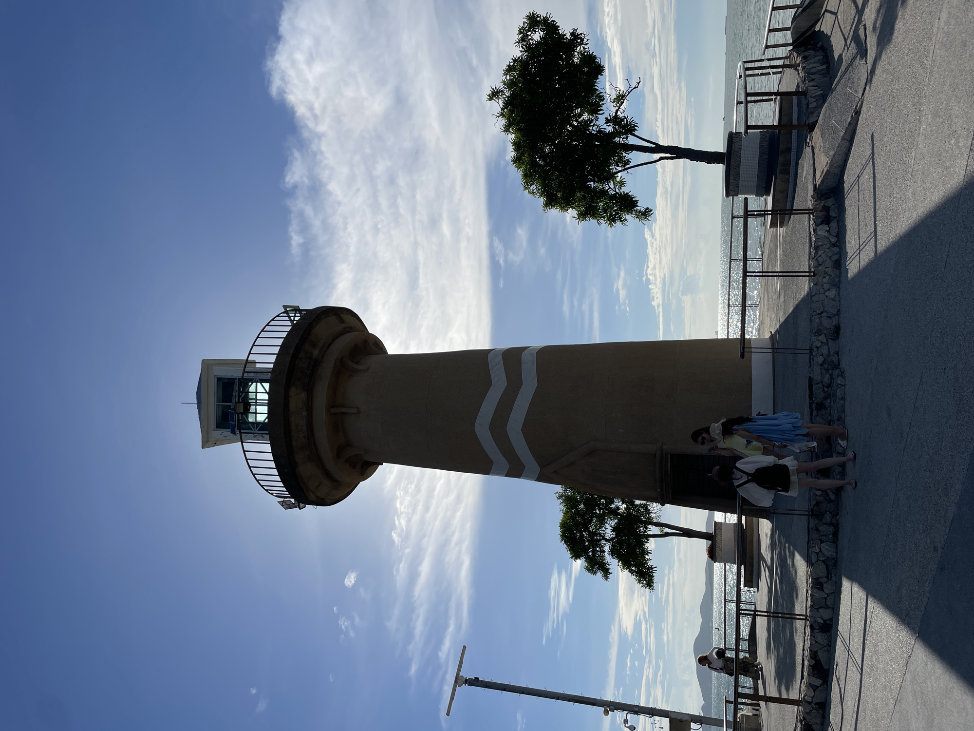 A light brown lighthouse with white wavy bands painted on it stands on a paved promenade by the sea in Pattaya City, Thailand. The lighthouse has a railing around its top and a lantern room. Two trees are planted near the lighthouse. Three women are standing and talking on the promenade in front of the lighthouse. One woman is standing by a railing on the left, looking out at the sea. The sea is calm with sunlight reflecting off its surface, and islands are visible in the distance. The sky is blue with scattered white clouds. A tall pole with a radar-like device is on the left side of the image. The scene is viewed from a low angle, with long shadows cast on the promenade.