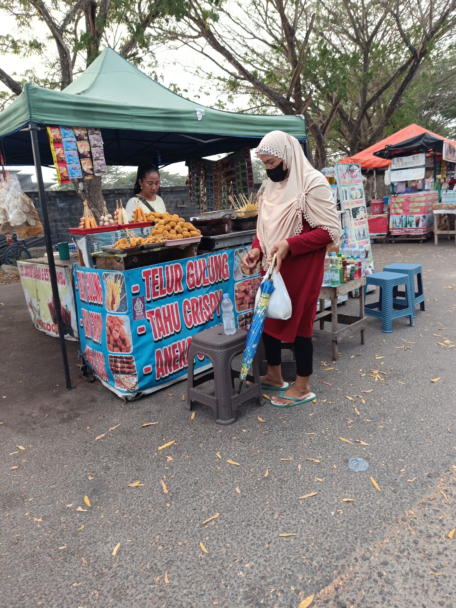 In the city of Kayu Agung, Indonesia, an outdoor street food stall displays an assortment of fried snacks, including egg rolls and crispy tofu, with a banner indicating "Telur Gulung Tahu Crispy Aneka". A vendor, an adult female with dark hair and a light-colored top, stands behind the stall, attending to the food. To the right, a customer, an adult female wearing a beige hijab, a red long-sleeved shirt, and black leggings, stands with a white plastic bag and a folded blue and yellow umbrella. She is wearing light green flip-flops. The street is paved with dark asphalt, scattered with fallen leaves. In the background, a second food stall with an orange canopy is visible, along with trees lining the street. The scene depicts a typical marketplace environment in Kayu Agung, with vendors and customers interacting amidst the offerings of local street food.