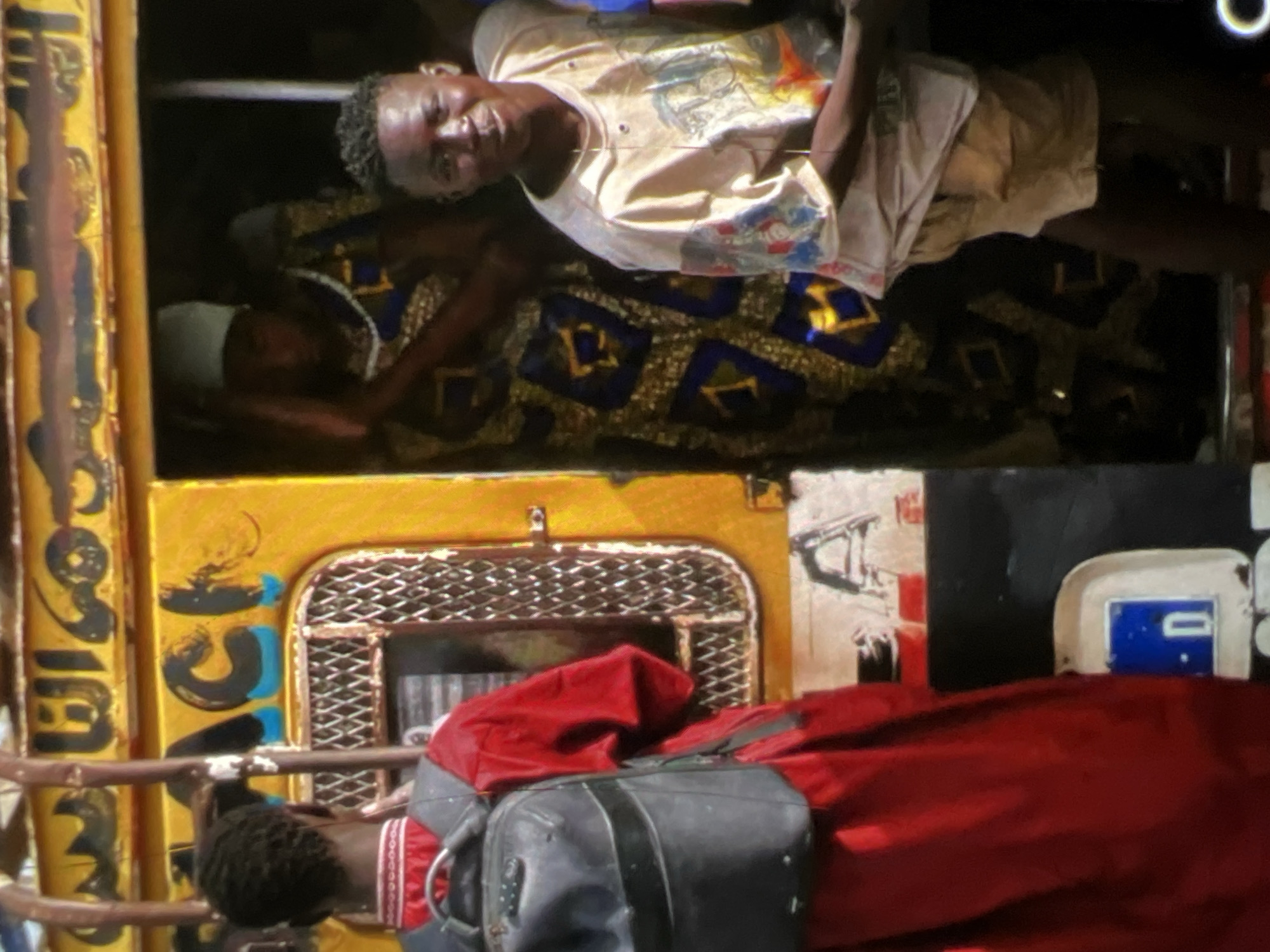 In Lagos, Nigeria, a vibrant yellow bus is partially visible, adorned with Arabic script along its upper edge and the word "ACH" in large, bold letters with blue accents. A young person in a red garment and a black backpack stands to the left, facing away from the camera. To the right, a person wearing a light-colored shirt with a graphic print and light-colored shorts smiles towards the viewer. Behind them, a person in a patterned blue and gold dress and a white head covering is visible within the bus. The scene suggests a moment of daily life and transportation in the bustling city.