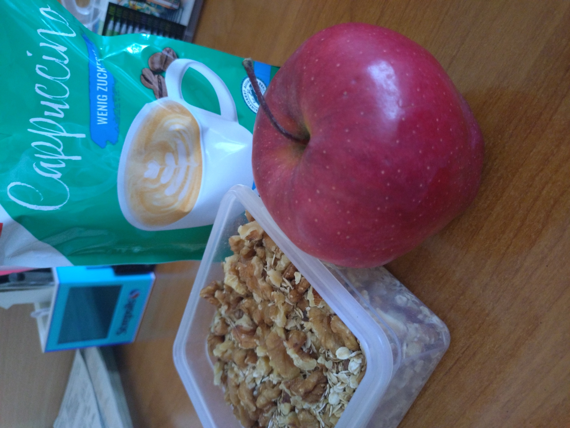 On a wooden surface, there is a red apple, a transparent plastic container filled with walnuts and oats, and a green package labeled "Cappuccino" with a picture of a cappuccino cup. A small, blue box with writing on it is partially visible in the lower-left corner of the image. The location is unknown.