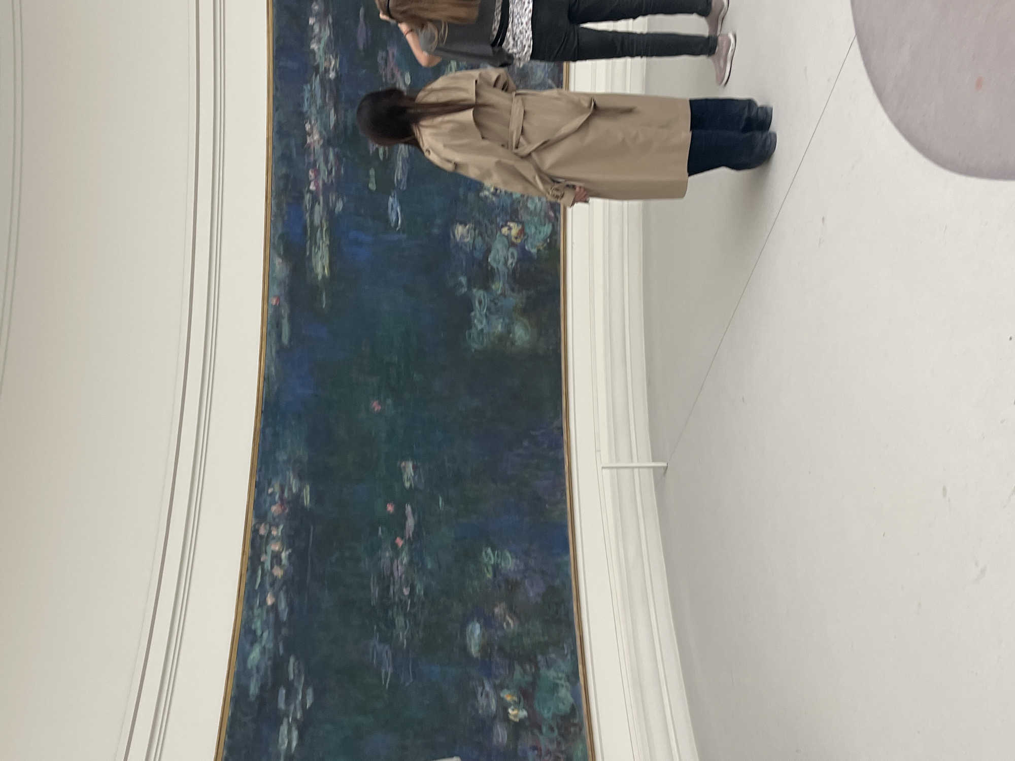 An indoor photograph, rotated ninety degrees clockwise, captures two individuals observing a monumental painting within a bright, minimalist gallery setting.
In the foreground, a person with long dark hair is seen from behind, wearing a light tan trench coat over dark trousers and shoes. Standing slightly to their left and further back, another individual, also viewed from behind, has lighter brown hair and wears a patterned grey and black top, dark pants, and light-colored sneakers.
The main subject is a vast, panoramic painting, identifiable by its style and scale as one of Claude Monet's "Water Lilies" (Nymphéas) series. The artwork, framed by a thin gold border, features rich blues, greens, and purples, with delicate touches of pink and yellow highlighting the lily pads and blossoms on the water. It extends along a subtly curved wall, creating an immersive experience for viewers.
The gallery space is characterized by pristine white walls and flooring, contributing to a serene atmosphere that focuses attention on the artwork. Architectural elements on the left side of the room (when upright) include vertical lines, suggesting the elegant, simple design of the building. A thin white rail or barrier is visible on the right, separating viewers from the artwork. The scene depicts a moment of quiet contemplation of art, indicative of a museum environment like the Musée de l'Orangerie in Paris, where Monet's "Water Lilies" are famously housed. No visible text is present.