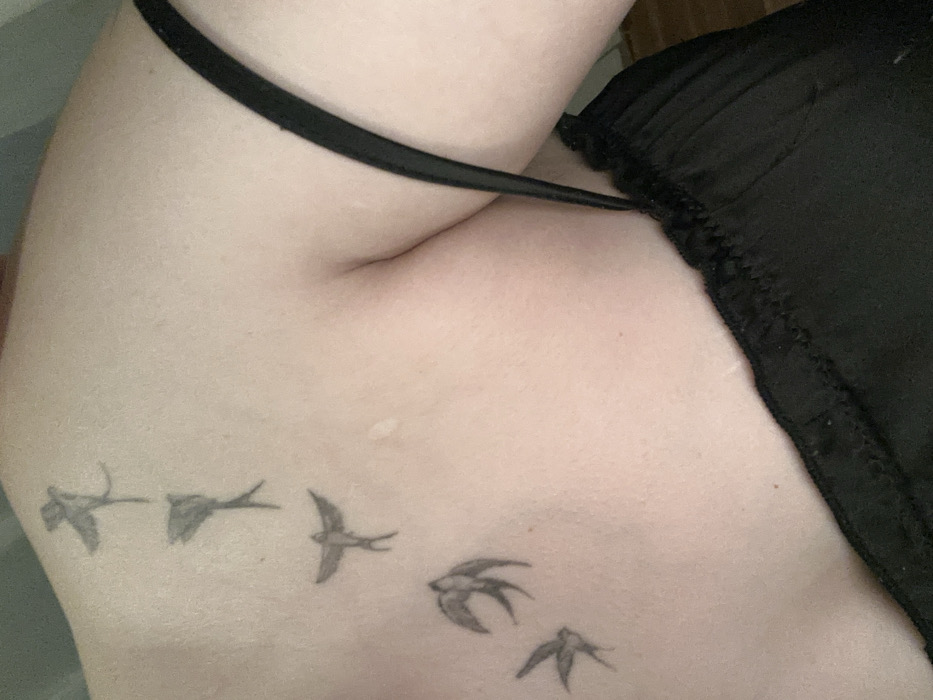The image displays a close-up view of a person's upper left torso and shoulder, specifically within the city of Chaikovsky, Russia. The individual's skin is light-toned. A series of five black-ink tattoos, depicting swallow-like birds in various stages of flight, are present along the left clavicle and upper chest area. The birds are arranged vertically, appearing to ascend from the lower chest towards the shoulder. The person is partially clothed in a black garment, visible on the right side of the frame, featuring a thin shoulder strap and a gathered or slightly textured top edge. A few subtle, raised skin imperfections or marks are discernible on the upper chest. The immediate background is indistinct, suggesting an indoor setting. No explicit actions or interactions are depicted.