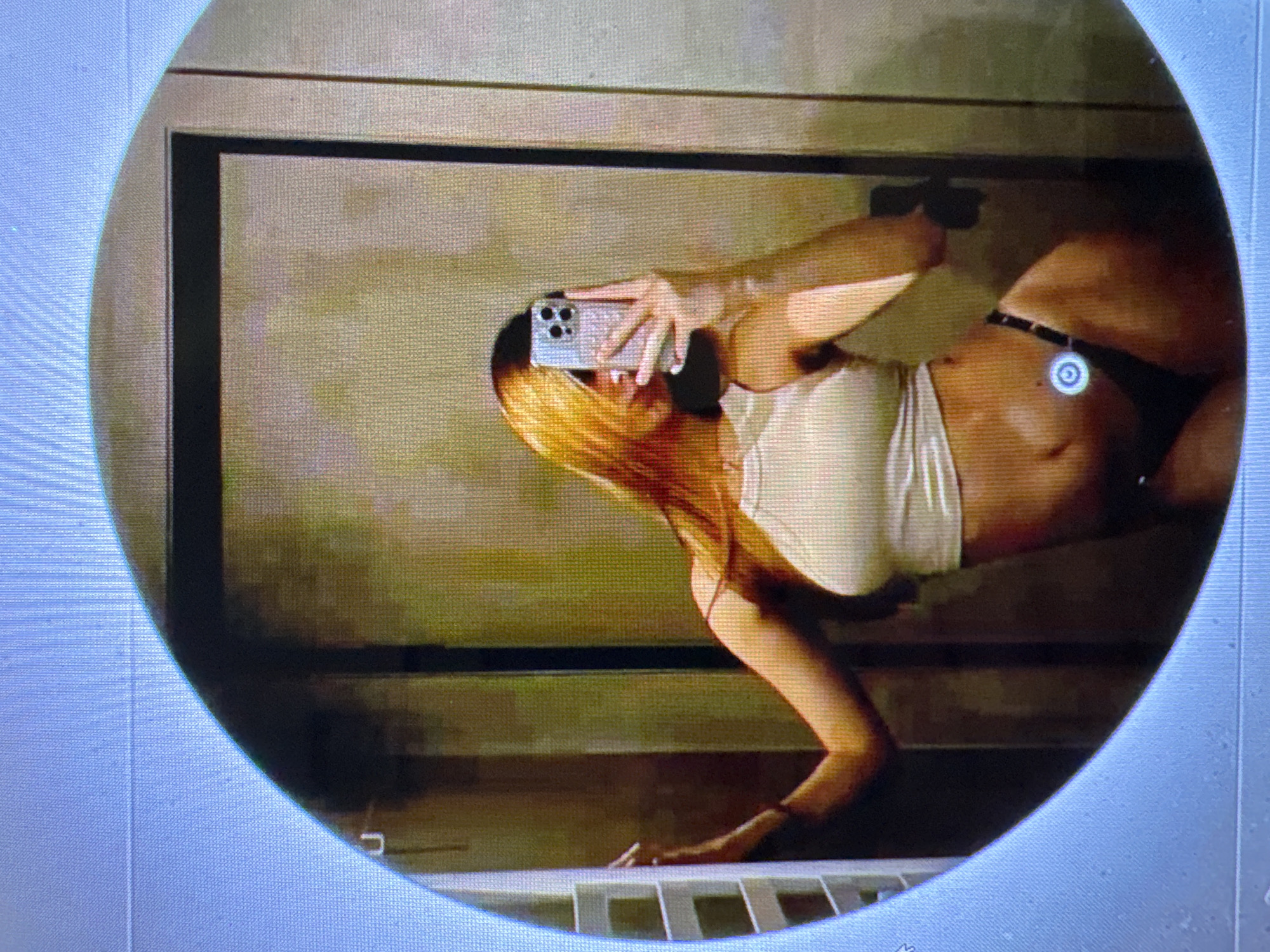 The image is a circular, pixelated photograph taken indoors, likely in a bathroom, as suggested by the presence of a towel rack on the left and a mirror reflecting the scene. A person, appearing to be a young woman with long, reddish-blonde hair, is taking a selfie in the mirror. She is wearing a light-colored, halter-neck crop top and dark-colored underwear. Her right arm is extended, holding a mobile phone, and her left hand is resting on the towel rack. The background is a textured wall, possibly tiled or paneled, in muted tones of beige and brown, with a dark vertical element which could be a doorframe or a cabinet. There is no indication of the city of Castelo Branco, Portugal, in the image itself.