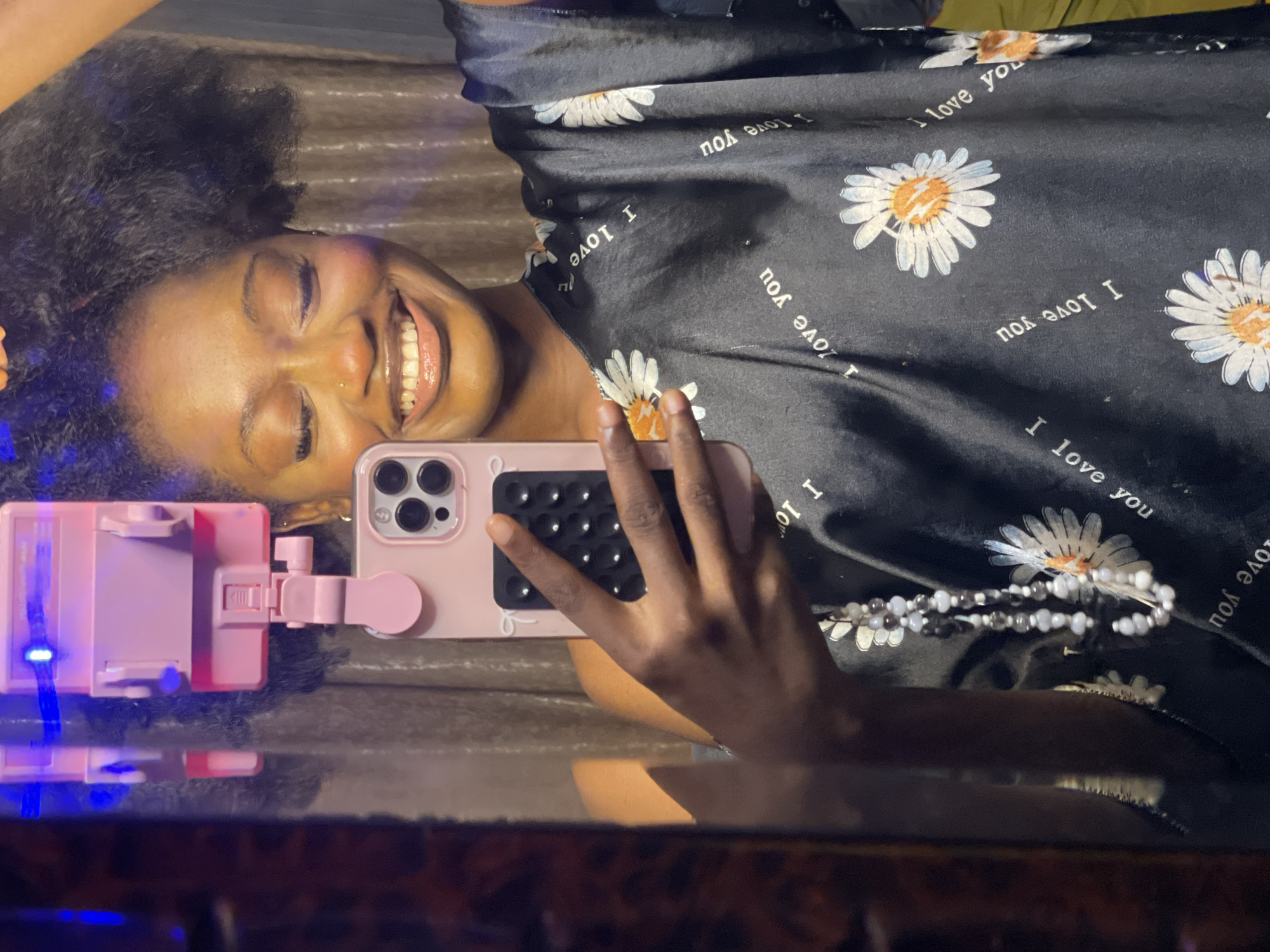 A young Black woman is taking a mirror selfie in what appears to be a bedroom. She is holding a pink smartphone in her right hand, which is angled to capture her reflection. The woman has dark, curly hair pulled up, closed eyes, and a wide smile. She is wearing a dark, silky garment with a floral print of white daisies and the phrase "I love you" printed repeatedly in white lettering. A beaded necklace with white, gray, and black beads hangs from her neck. The background consists of a brown, patterned curtain and a pink phone stand with blue lights attached to a mirror. The lighting is dim, with some blue light casting reflections on the mirror and curtain. The overall impression is of a casual, personal moment. There are no discernible Ado, Nigeria specific elements.