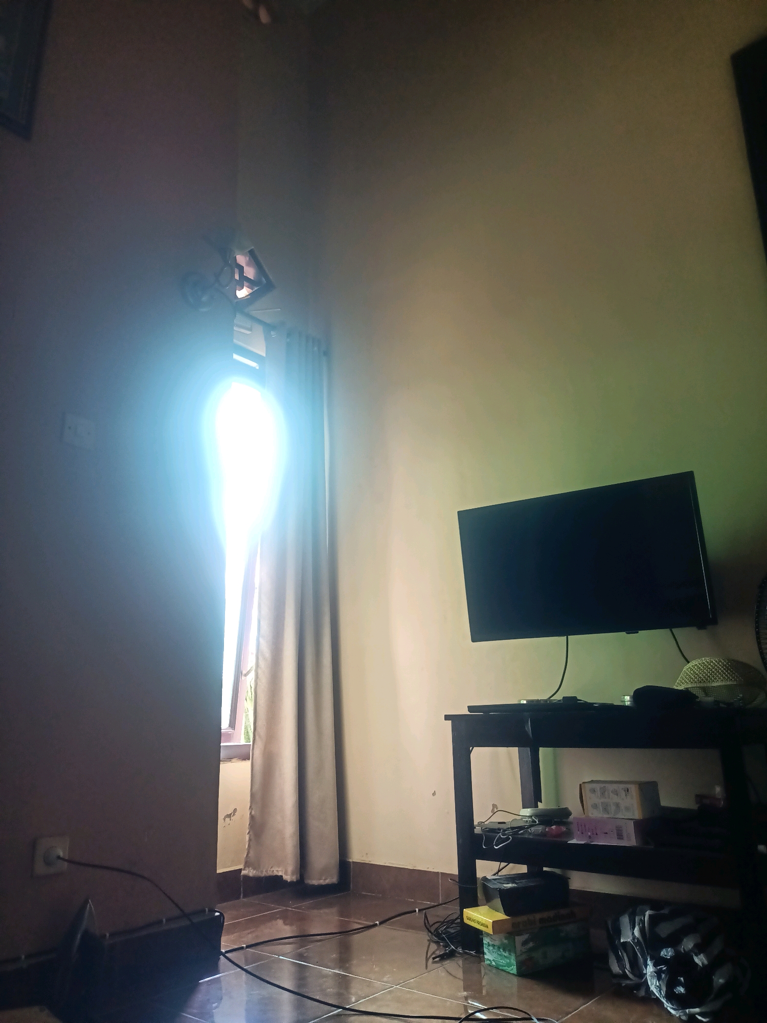 The image shows an indoor scene, likely a living room, with a window letting in bright sunlight. A beige curtain is partially drawn, obscuring the view outside. To the right of the window, a black television is mounted on the wall above a black entertainment stand. The stand holds several boxes, cables, and a white, textured hat. The floor appears to be tiled and is reflective, showing the lighting from the window. A power outlet with a black cable plugged into it is visible on the wall to the left of the window. The room has beige walls. The bright light from the window suggests it is daytime. The scene does not provide any specific visual cues to pinpoint its location within the city of Trenggalek, Indonesia.