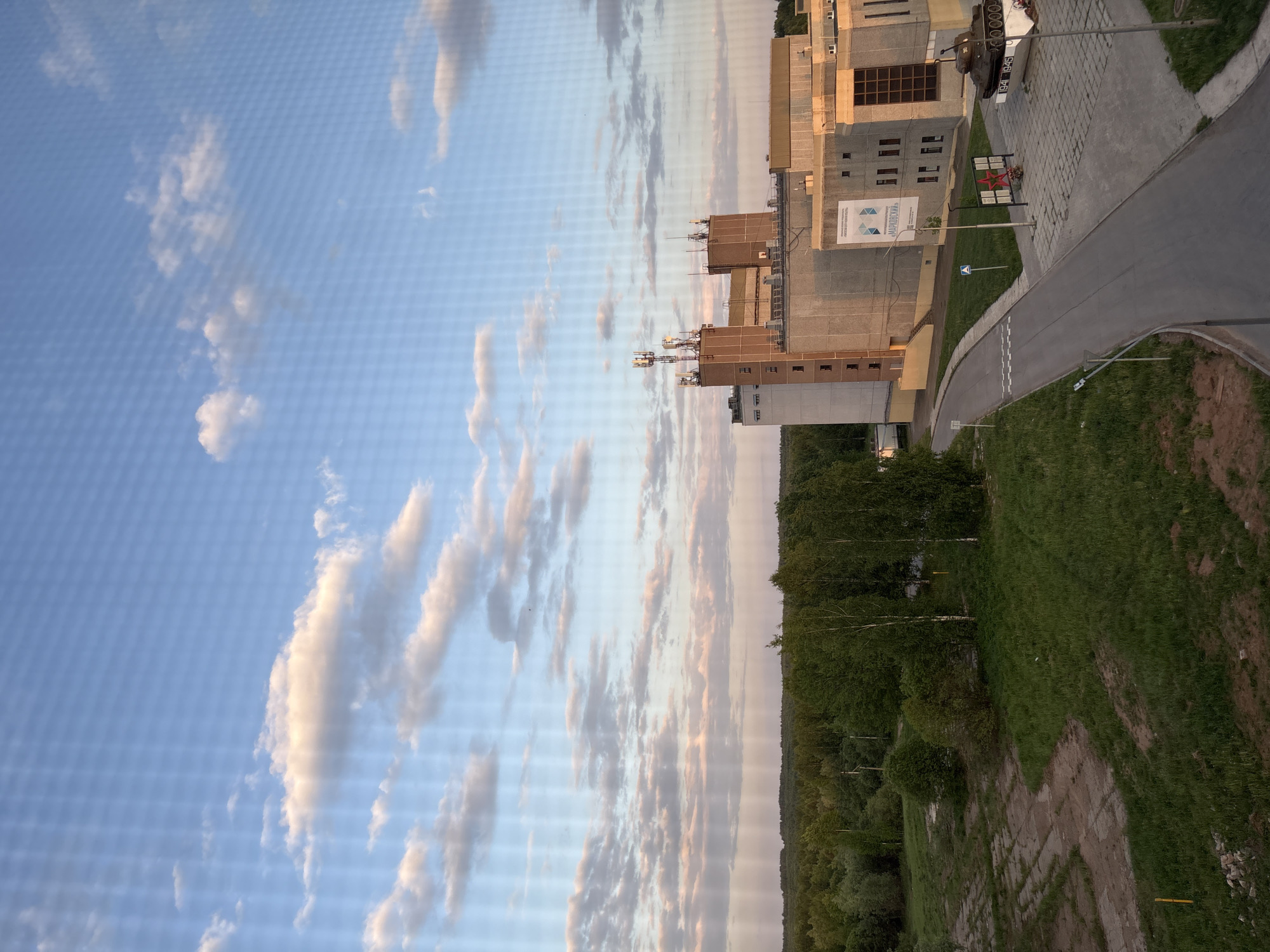The image shows a landscape in Марковский, Russia. On the right, there is a large, multi-story concrete building with a series of antennas on top. Adjacent to the building is a road leading to a monument consisting of a tank and a star-shaped memorial. The road is lined with grassy areas. Beyond the road, a dense forest stretches into the distance. The left side of the image is dominated by a blue sky filled with numerous white clouds, suggesting a partially cloudy day. The image appears to be taken from an elevated vantage point, providing a panoramic view of the urban and natural landscape.
