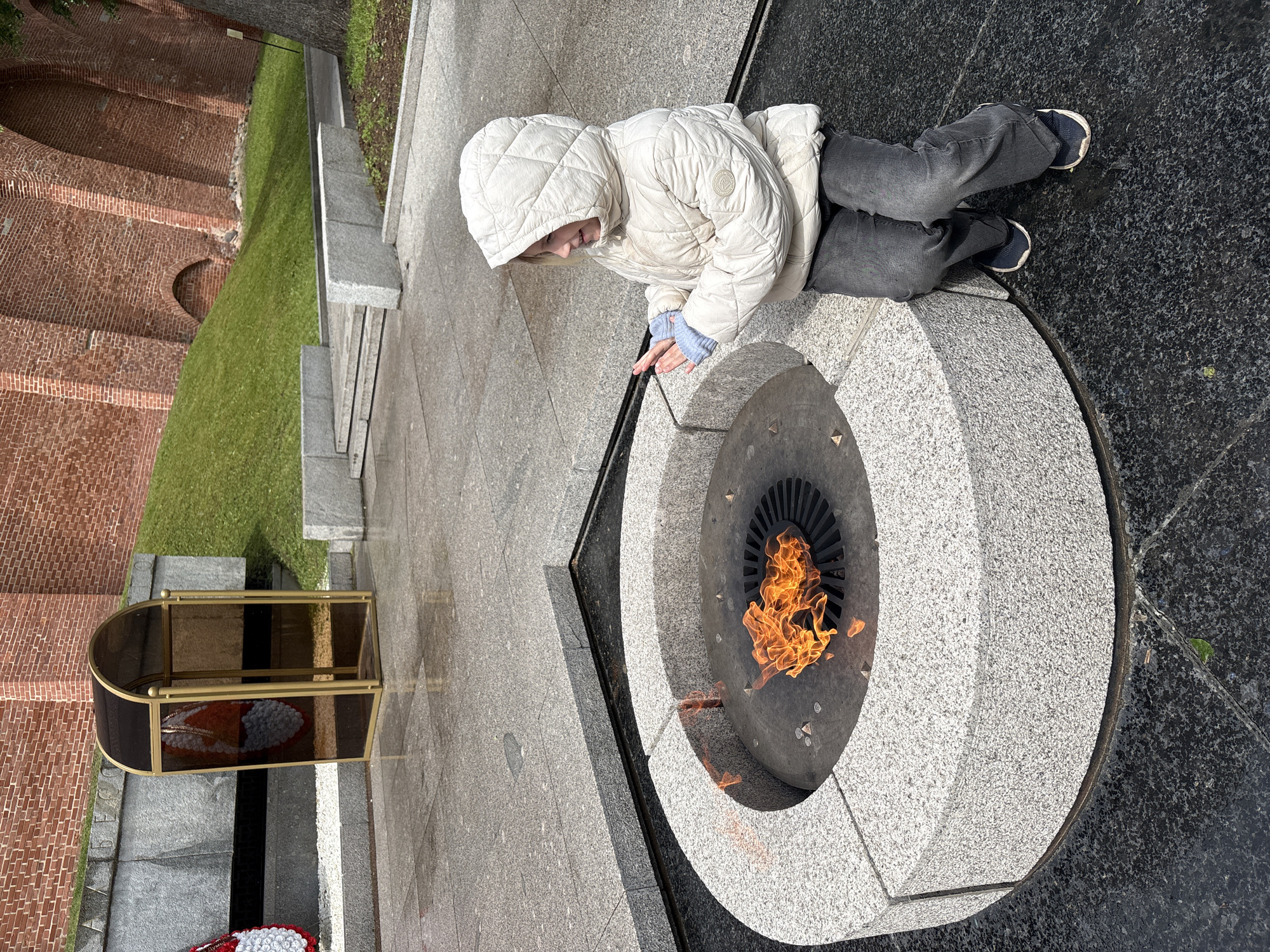 A person in a white quilted jacket with the hood up is leaning over a circular Eternal Flame monument at the Kremlin in Veliky Novgorod, Russia. The person is wearing grey pants and dark shoes. The flame burns in the center of the monument. A glass case with a floral wreath is behind the flame. Brick walls and grassy embankments are in the background.