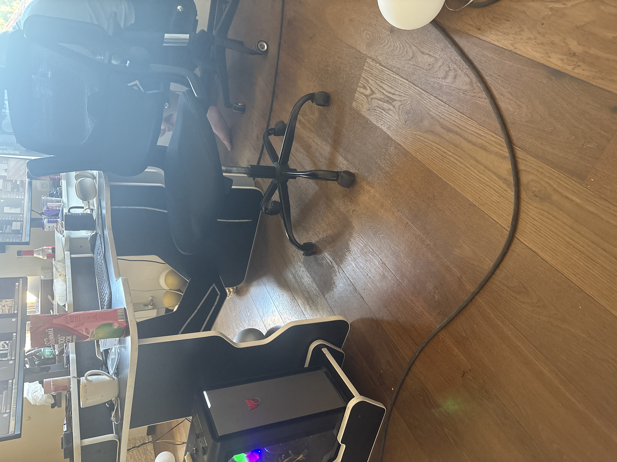 A computer desk with dual monitors is visible in the background. On the desk, there is a white mug, a box of juice, and a keyboard. In the foreground, a black mesh office chair is positioned to the right of the desk. A computer tower with green and purple LED lights is located to the left of the desk. A black cable runs across the wooden floor. The scene appears to be an indoor workspace.