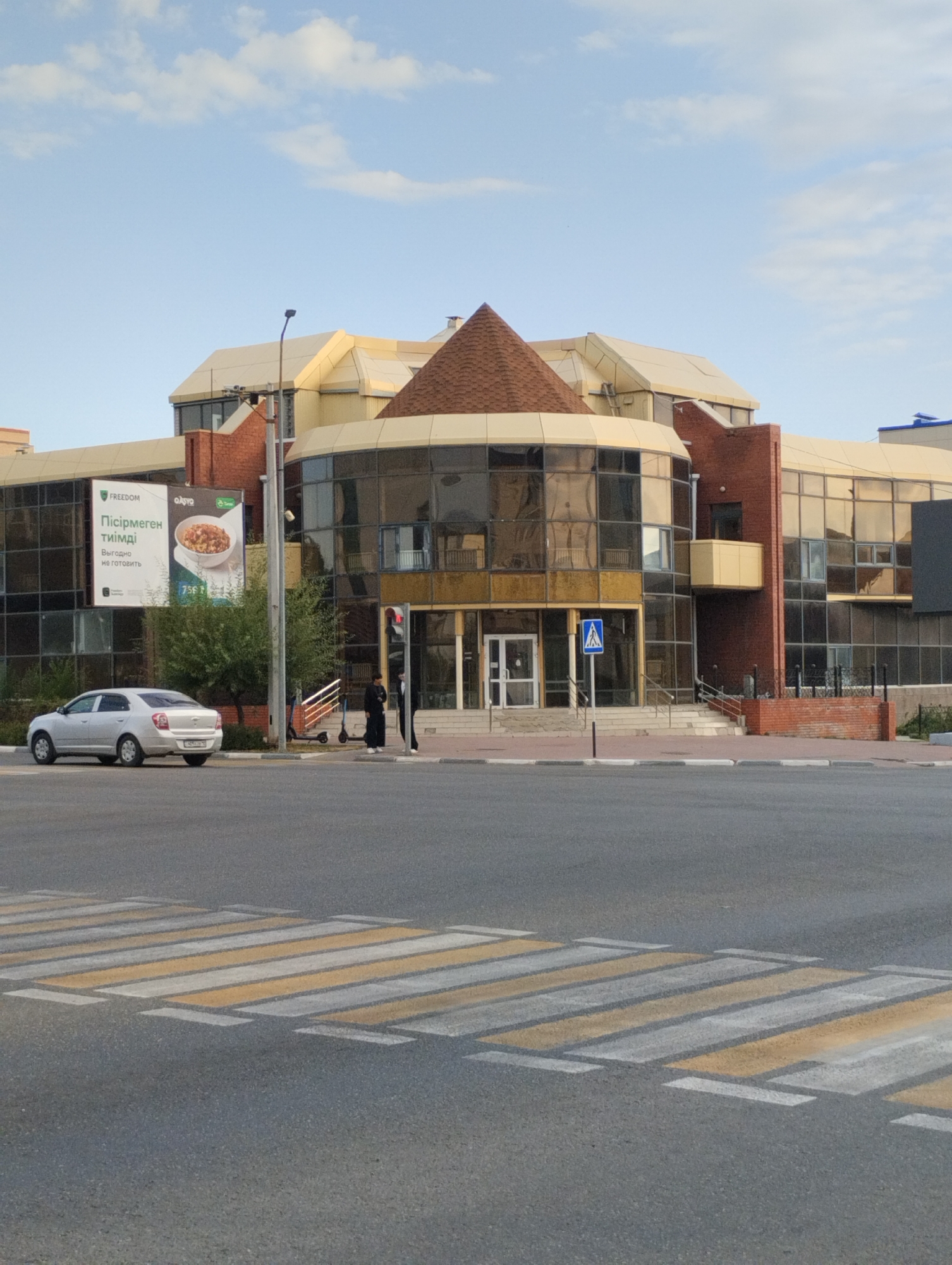 This image, taken in Kostanay, Kazakhstan, captures a modern commercial building with extensive glass facades and an unusual conical roof element. In the foreground, a pedestrian crossing is visible on an asphalt road. A silver sedan is parked on the left side of the road, facing away from the viewer. To the right of the car, a tall street lamp and a traffic signal are present. Two individuals, one wearing a dark outfit and the other in white, are standing near the building's entrance. Two electric scooters are parked on the sidewalk. A large billboard advertising "FREEDOM" and a food item is mounted on the building's exterior. The sky above is partly cloudy.