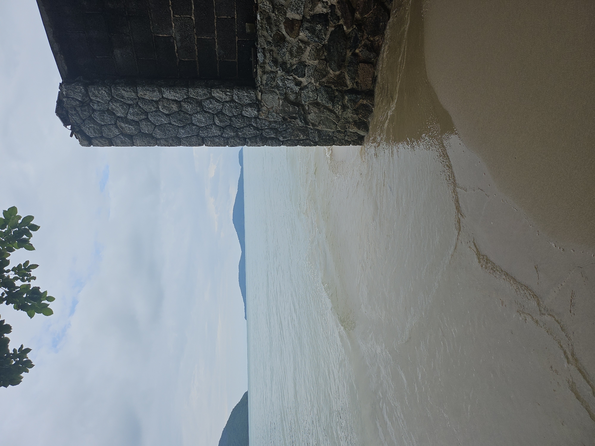 The image depicts a waterfront scene in Rawai, Thailand. On the right side of the frame is a stone structure with a weathered, dark grey finish. The stone is laid in a hexagonal pattern, forming a textured wall. Adjacent to this stone section, the wall transitions to dark grey brickwork. The structure appears to be a building or a retaining wall situated at the edge of the water.
The sea is calm with gentle waves lapping onto a sandy shore. The water is a muted blue-grey, reflecting the overcast sky. In the distance, several islands with rounded, green-covered hills are visible on the horizon. The sky is cloudy with patches of light blue. Overhead, on the left, a cluster of green leaves from a tree hangs into the frame, partially obscuring the sky. The foreground consists of wet sand where the water has receded. No people are visible in the image.