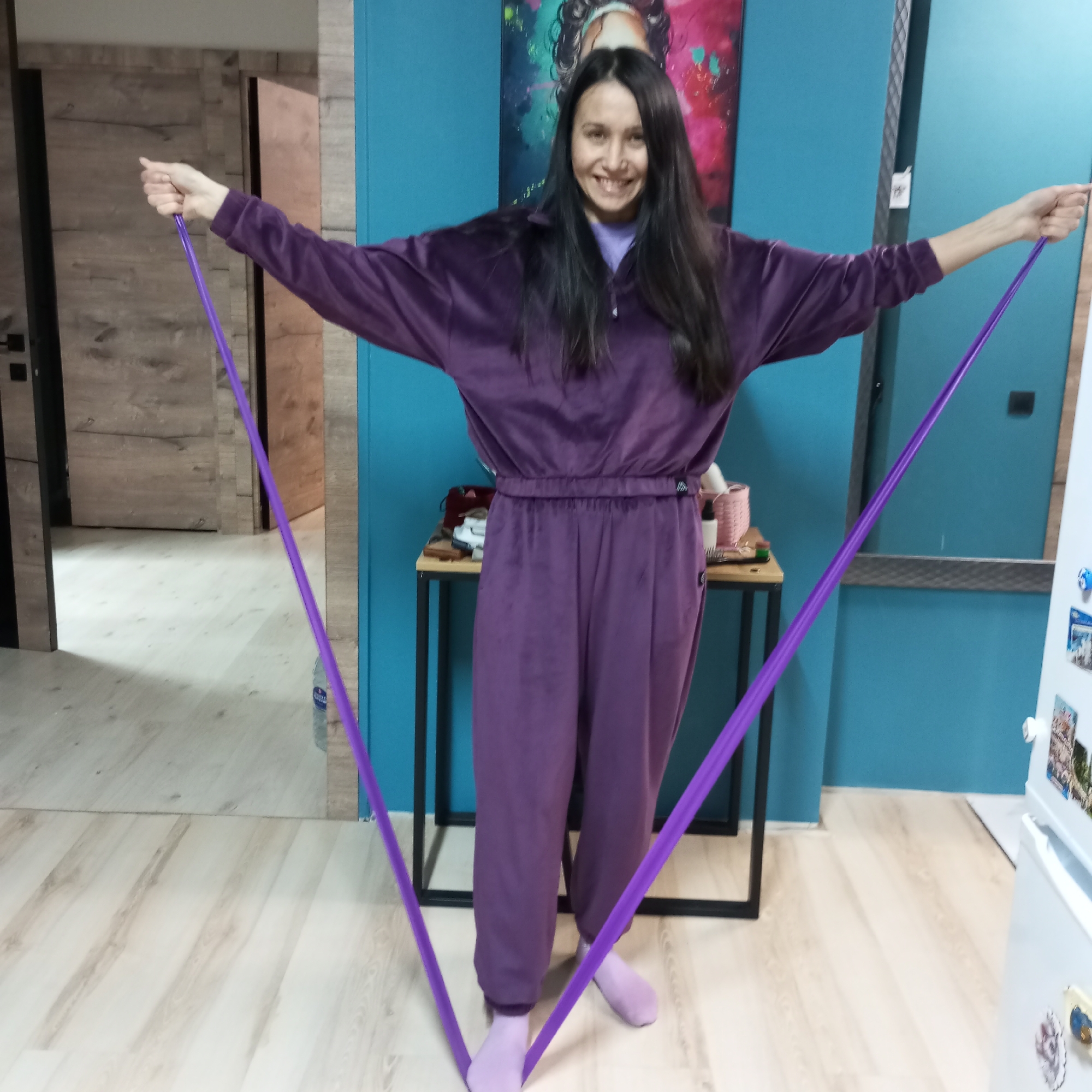 A woman stands indoors holding a purple exercise band. She wears a purple velour tracksuit and matching socks. She smiles and has long, dark hair. The exercise band is taut in her hands, running down to the floor and under her feet.
Behind her is a teal-painted wall with an abstract painting. A small table with items on it is in front of the wall. To the left is a wooden door leading to another room. To the right is a white refrigerator with magnets. The floor is light wood-look laminate.
There is no indication this image was taken in the city of Pervomaysky, Russia.