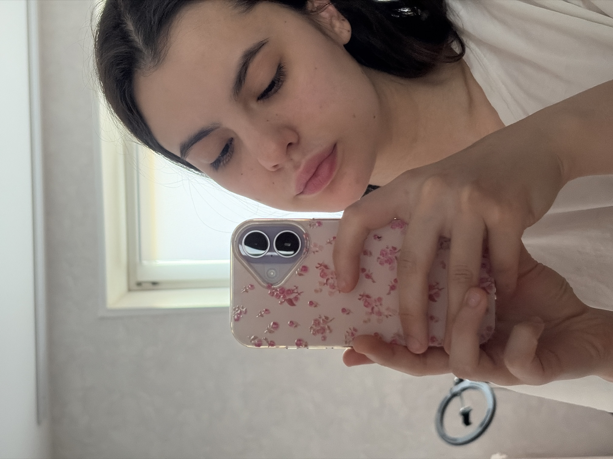 A young woman is taking a selfie with a phone in a bright indoor space, possibly a bathroom. She has dark hair, fair skin, and is wearing a white shirt. The phone case has a light pink background with a floral design. Part of a window and a gray patterned wall are visible. There is a key on a keyring hanging near the bottom of the image. Location information suggests Chaikovsky, Russia, but this is not explicitly visible in the image.