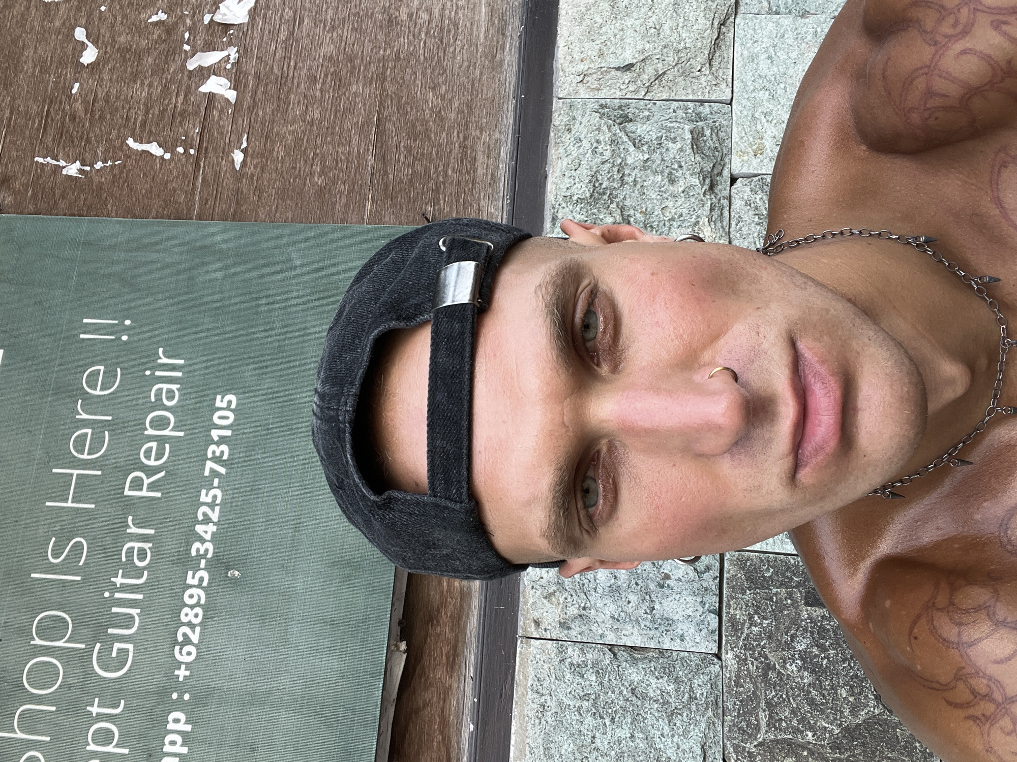 In North Kuta, Indonesia, a young adult male with tanned skin and multiple tattoos on his shoulders is captured in a close-up selfie. He wears a dark baseball cap and a silver chain necklace with pointed pendants. A silver hoop earring is visible in his left ear, and a gold nose ring adorns his right nostril. His eyes are a striking green, and he has rosy cheeks. The background consists of a rough stone wall and a wooden surface with peeling white paint, suggesting an outdoor setting, possibly near a business that offers guitar repair services, as indicated by text on a green sign in the upper left corner of the image.