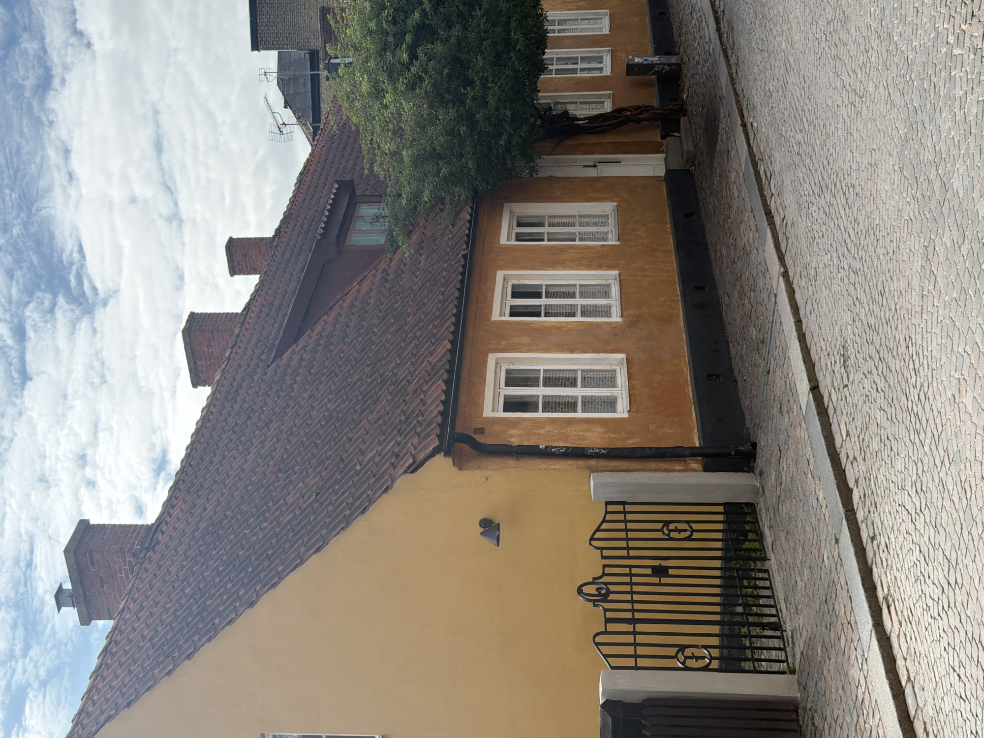 The image depicts a narrow cobblestone street in Lund, Sweden, with a two-story building dominating the scene. The building features an orange-yellow facade and a red-tiled roof with multiple chimneys. Several white-framed windows are visible on the building's facade. A black wrought-iron gate with decorative circular elements is positioned in front of a portion of the building, bordered by white posts and a wooden fence. A large, leafy tree grows near the side of the building, partially obscuring the entrance. The sky above is cloudy, with a mix of blue sky and white clouds. The street surface is made of evenly laid cobblestones, with a raised concrete curb running along its edge. The overall impression is of a historic European streetscape.