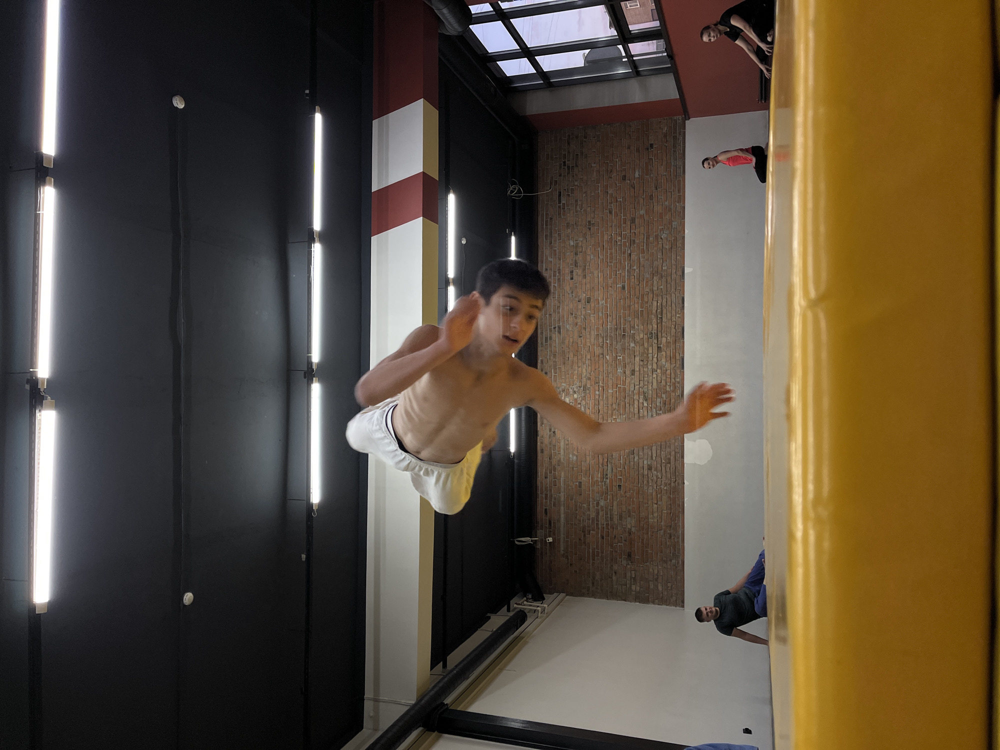 A young man is suspended mid-air in a sideways shot taken indoors. He appears to be performing an acrobatic or gymnastic maneuver, with his body almost parallel to the ground. He is bare-chested and wearing white shorts.
The setting seems to be a gymnasium or an indoor sports facility. The environment includes black and white vertical panels with fluorescent lights, brick wall, a clear ceiling, and other figures that appears to be observers in the distance and near a yellow pillar.
The location might be in Krasnoyarsk, Russia, but there is no element that specifies it directly.