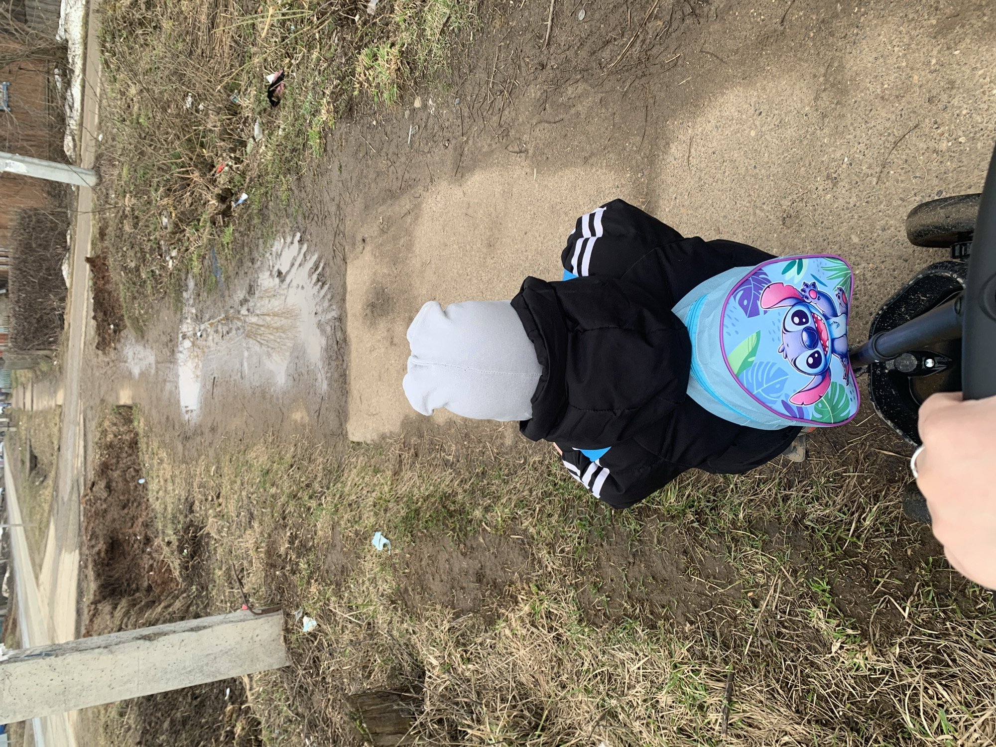 A child sits on a tricycle on a dirt path in Фоки, Russia. The child wears a light gray knit hat and a black jacket with white stripes on the sleeves. A light blue backpack with a "Lilo & Stitch" character print is attached to the tricycle. The path is muddy and edged with dead grass, weeds, and scattered debris, including what appears to be discarded medical masks. Water has pooled in some spots on the path. A utility pole and buildings are visible in the background. A hand is visible holding the handle of the tricycle.