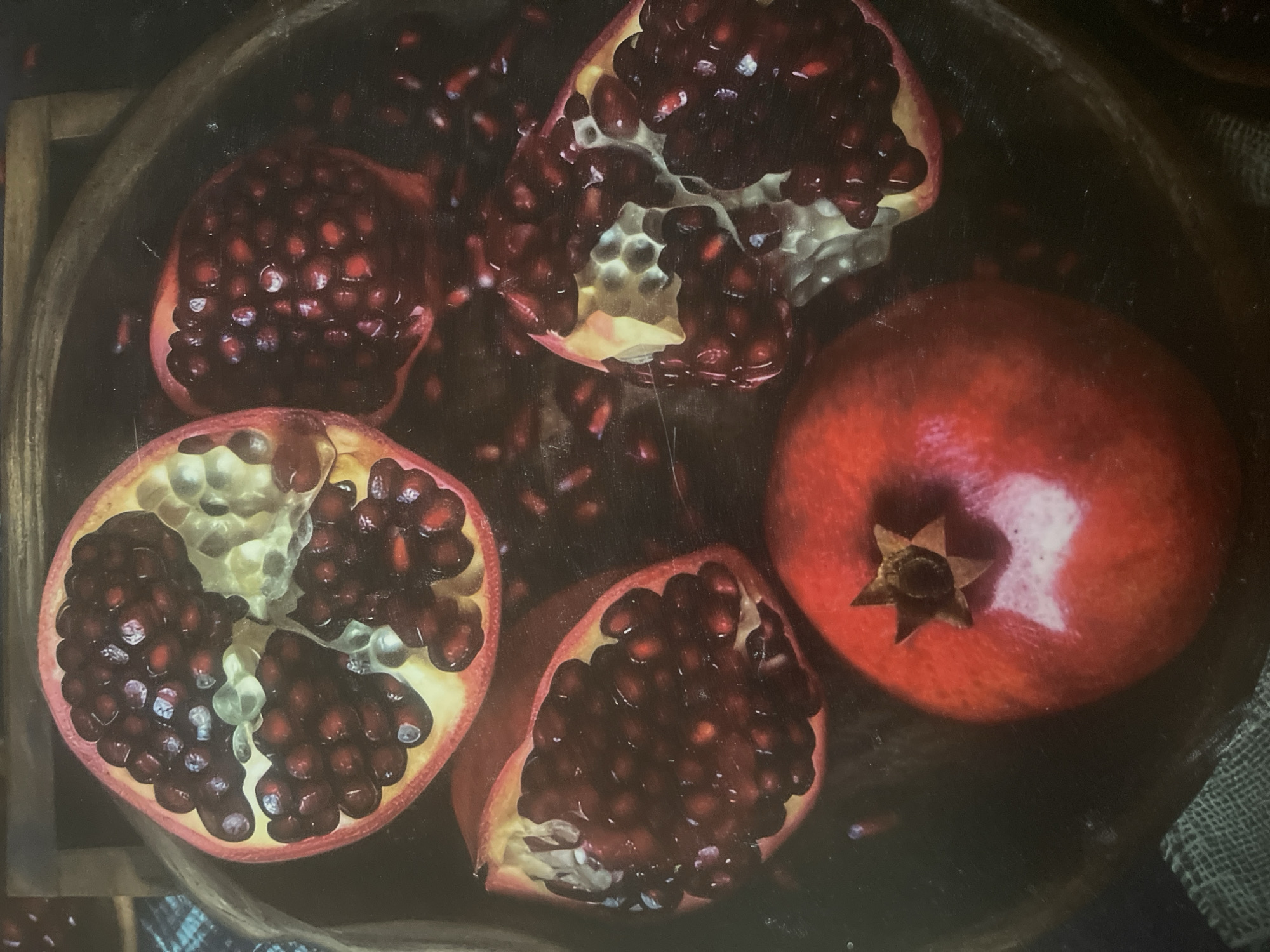 A close-up, overhead shot displays a collection of pomegranates, some whole and some cut open, arranged in a wooden bowl. The vibrant red fruit contrasts with the dark background, with arils of the cut pomegranates spilling out. The image is dark and moody, with dramatic lighting that highlights the textures of the fruit. The scene is devoid of any human presence and does not contain any elements that would suggest a specific location within Minsk, Belarus.