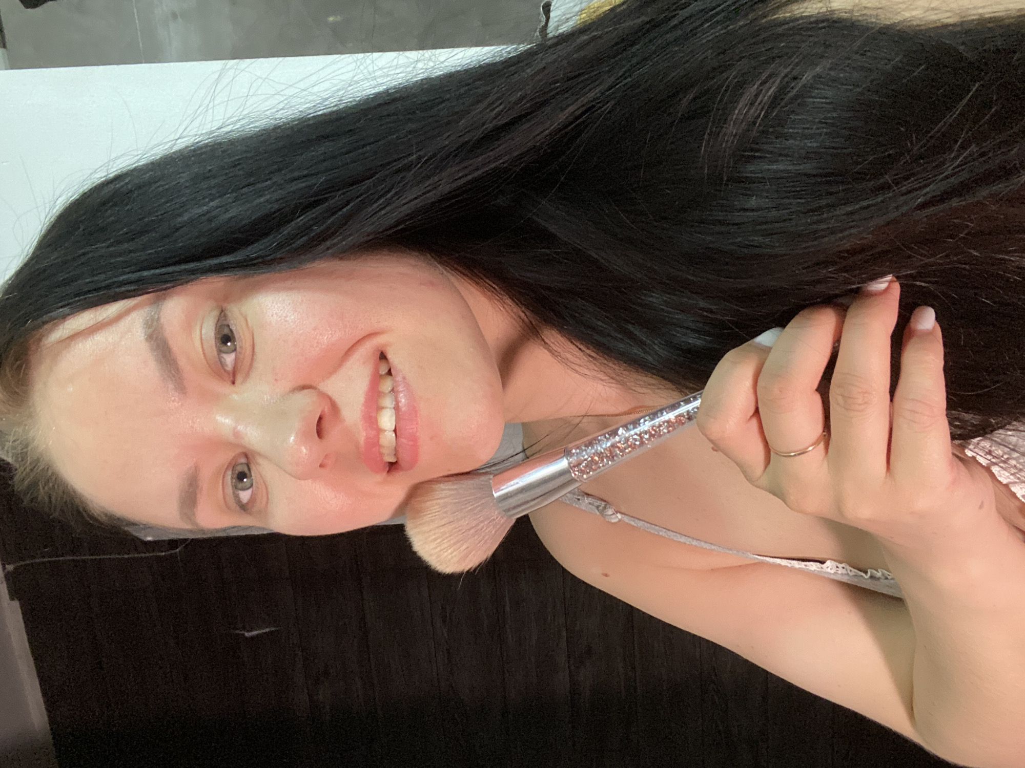 A woman with long dark hair smiles while holding a makeup brush to her face. She is wearing a light-colored top with lace detailing. Her left hand is visible, adorned with a gold ring. The background appears to be a combination of a white wall and a darker, possibly wooden, surface. There is no indication of the image's location in Chaikovsky, Russia.