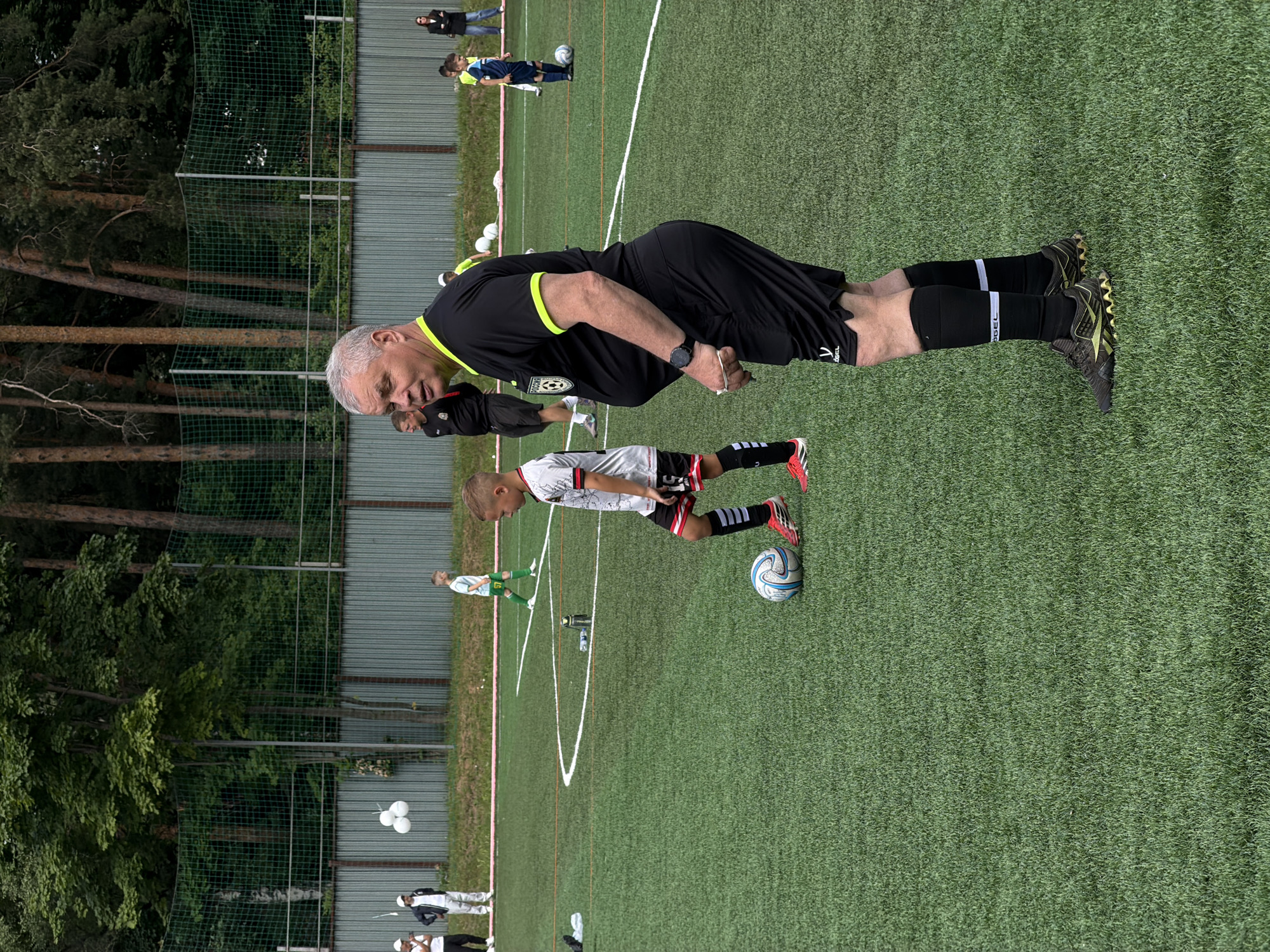 A group of people are on a green artificial turf soccer field. The scene is likely set in Bykovo, Russia, given the context. There are several individuals, including a man in a black soccer referee uniform with yellow trim, who appears to be addressing a young boy in a white and red soccer uniform. The boy is standing next to a soccer ball. Other individuals, some appearing to be younger players in soccer uniforms, are visible in the background on the field. The background also shows a metal fence and trees, suggesting an outdoor setting. The time of day appears to be daytime, with bright sunlight casting shadows, indicating clear weather. White balloons are visible in the background near the fence. There is some text visible on the referee's uniform, including a circular emblem with a soccer ball and the word "REGEL" on the side of his shorts. The overall atmosphere seems to be one of a soccer practice or game.