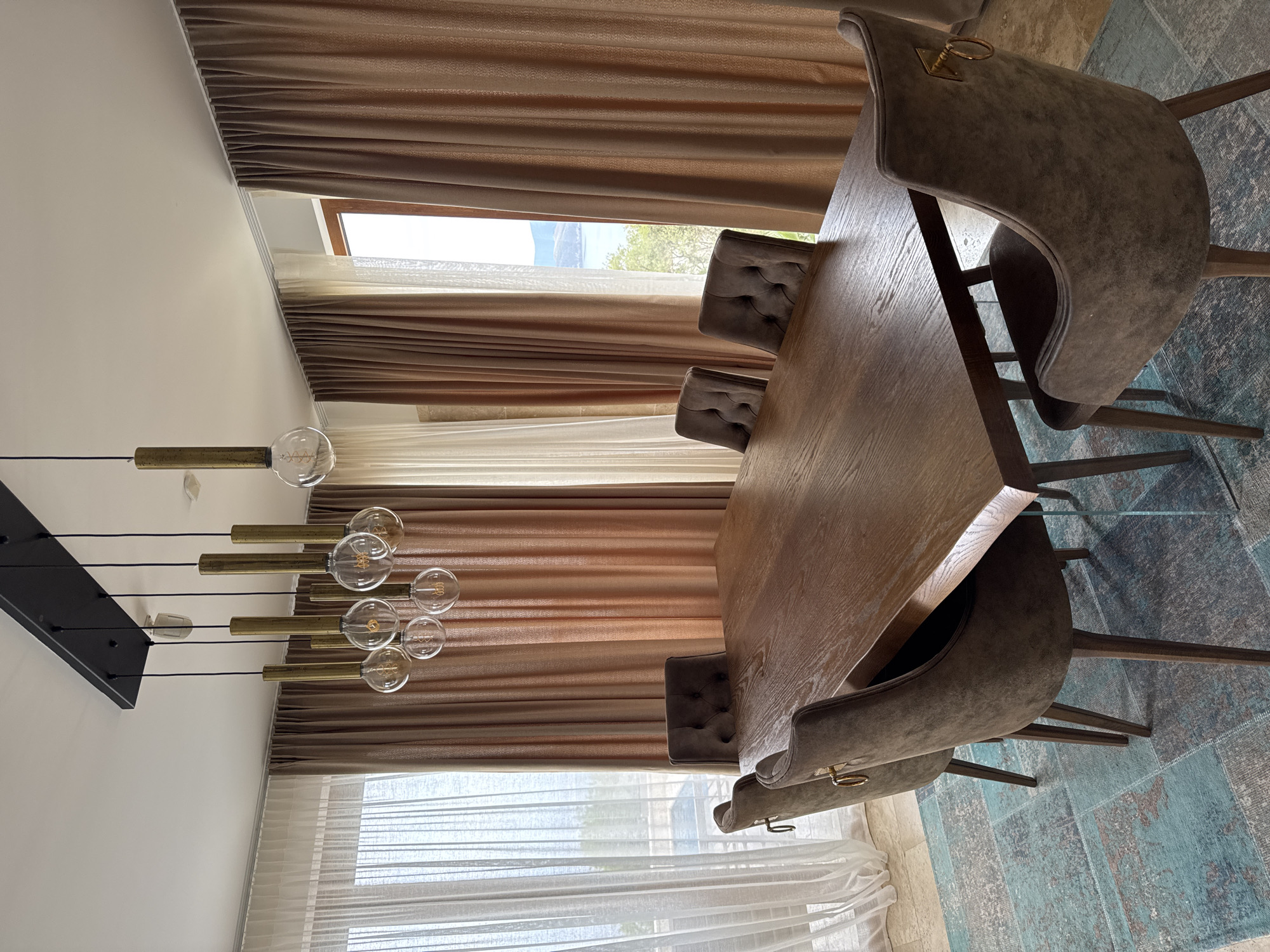 Here is a description of the image:
The image depicts a dining room setting. A large wooden table is the focal point, surrounded by upholstered chairs. The room is illuminated by a modern chandelier with multiple exposed bulbs. Large windows are covered by layered curtains, offering a glimpse of an outdoor landscape with trees and mountains. The scene is located in Blizikuće, Montenegro, a coastal town with a mountain backdrop. The overall aesthetic suggests a contemporary and stylish interior design.