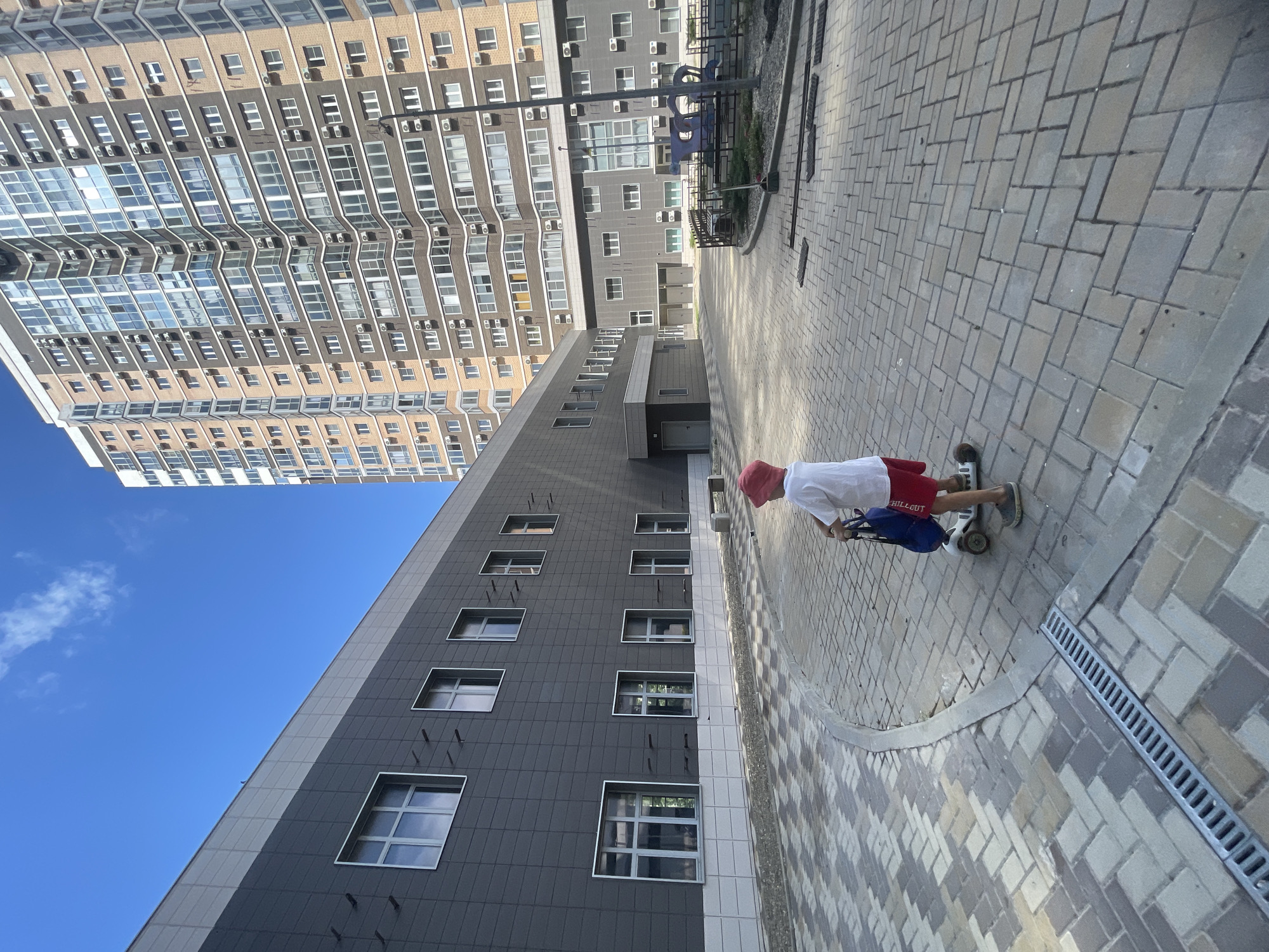 The image depicts a scene in the city of Novorossiysk, Russia. A young child wearing a red hat, white t-shirt, and red shorts is standing on a scooter with a blue bag attached to the handlebars. The child is positioned on a paved courtyard area with a distinctive pattern of gray and beige interlocking pavers. To the left, a modern building with a dark gray facade and numerous windows is visible. Further in the background, a tall, multi-story residential building with a brick and glass facade dominates the skyline. The sky is clear and blue with a few wispy clouds. The scene appears to be outdoors during the daytime.