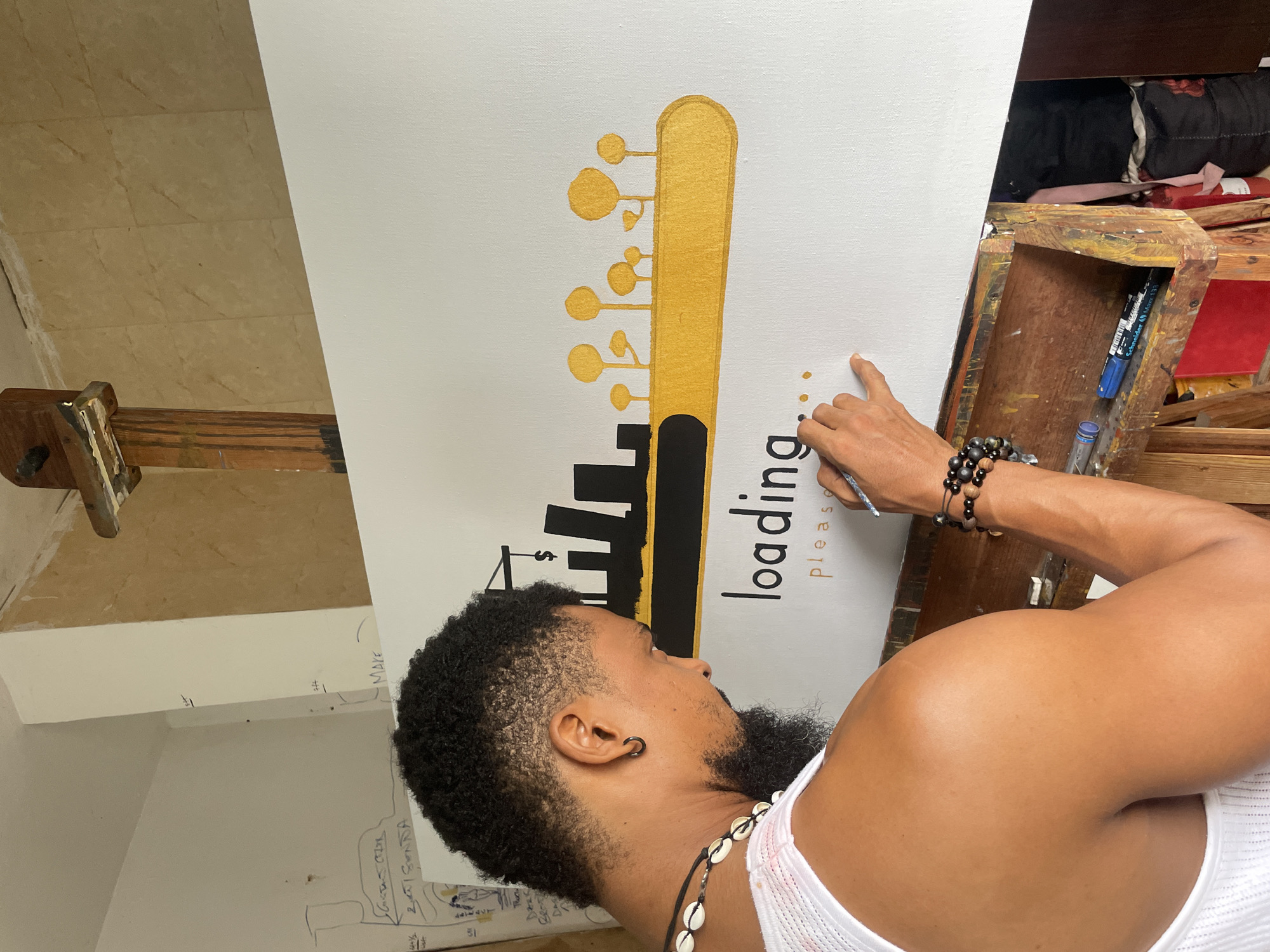 A man is painting a picture of a guitar headstock on a white canvas. The man has dark curly hair and a beard, and he is wearing a white tank top and bracelets on his left wrist. The painting features a gold guitar headstock with black skyscraper silhouettes and the words "loading... please". The painting is on an easel in a room with beige tiled floors and white walls. There is a wooden art supply box with paintbrushes and markers on the right side of the frame, and the wall to the left is covered in sketches and notes. This picture was taken in Ado, Nigeria.