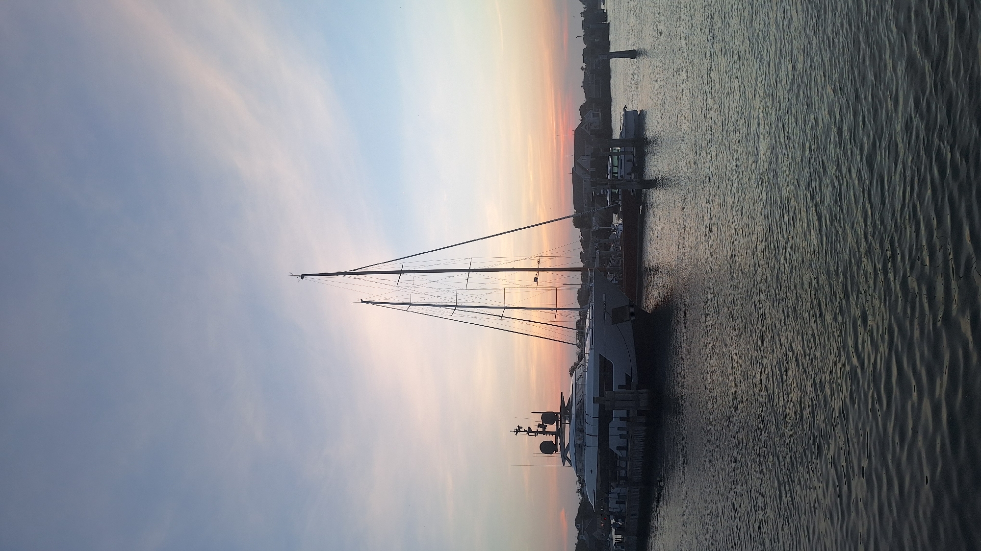 The image depicts a harbor scene at dusk in Nantucket, Massachusetts, United States of America. Dominating the foreground is the calm, rippling water of the harbor. Moored along a dock in the mid-ground are several vessels. The most prominent is a large, modern white yacht with a sleek profile and multiple decks and radar equipment. To its right, a tall-masted sailboat stands silhouetted against the sky. Behind these vessels, smaller boats and a cluster of buildings are visible along the shoreline, suggesting a residential or commercial area. The sky above is a gradient of soft blues and pinks, indicative of a sunset or sunrise, with wispy clouds. The overall atmosphere is peaceful and serene, capturing a moment of quietude in the harbor.