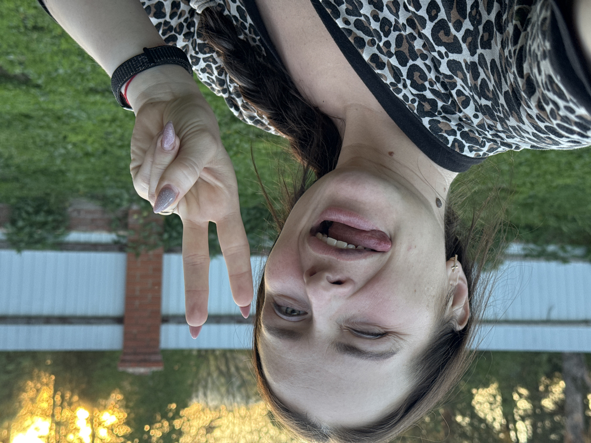 A woman is positioned upside down in front of an outdoor scene that includes a white fence, brick pillar, green grass, and trees. The background suggests either a rural or suburban environment.
The woman is wearing a leopard-print top with a black collar. She has a dark-colored hair and a bracelet on her left wrist. The woman is sticking her tongue out and winking. She is holding up her left hand, making a peace sign with her fingers.
No specific details in the image suggest that this photo was taken in Rusalevka, Russia.