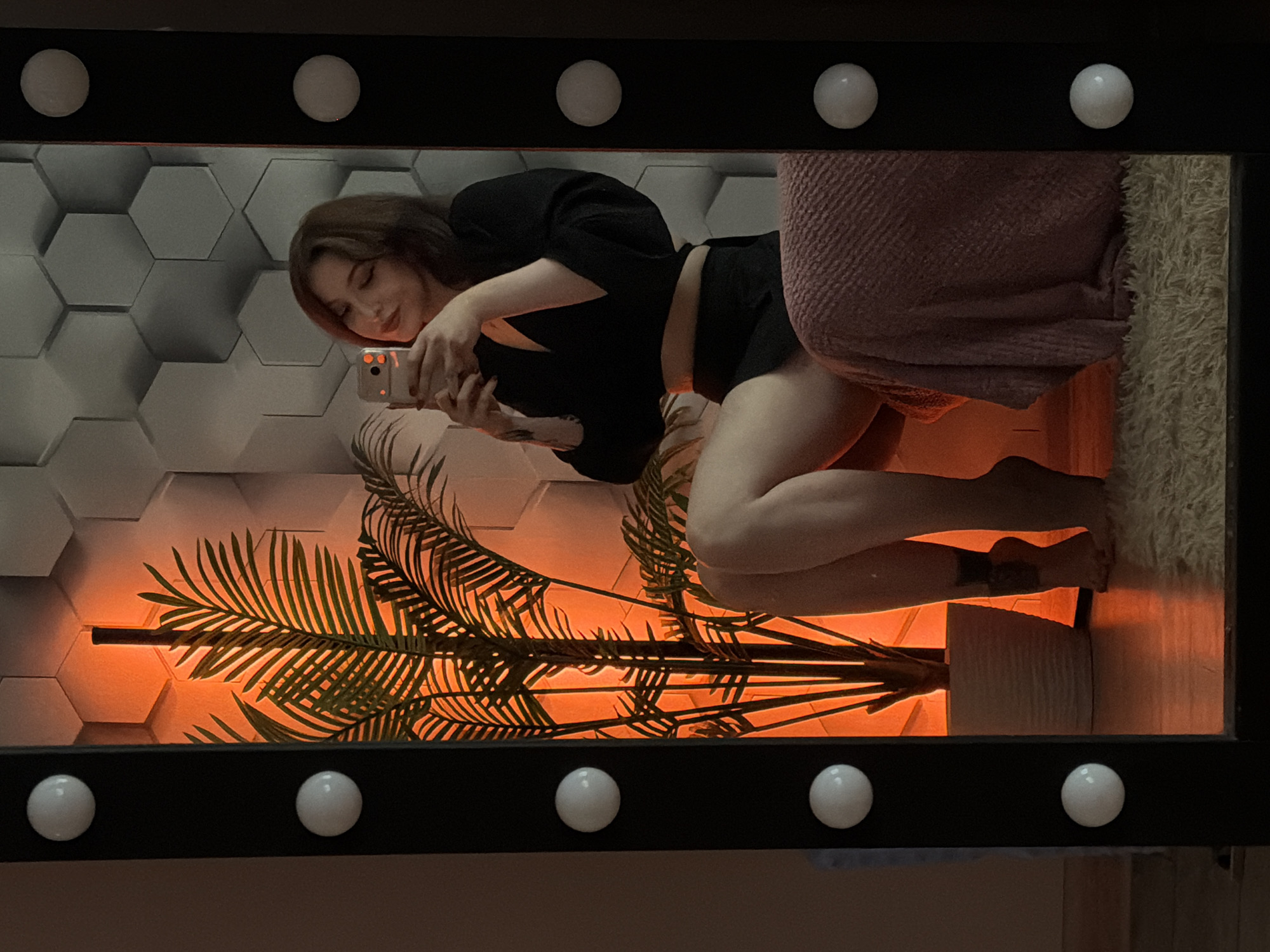 A young woman with shoulder-length brown hair, wearing a black crop top and black shorts, takes a selfie in a large vanity mirror. She is sitting on a textured pink blanket, with a potted palm plant to her left, illuminated by an orange light. The background wall is made of hexagonal panels. The mirror frame has a black border and is adorned with evenly spaced white light bulbs. The woman's right leg is bent, her foot resting on the floor, and a tattoo is visible on her ankle. The floor is covered with a cream-colored shaggy rug. The scene is indoors, with no visible indication of a specific city or location.