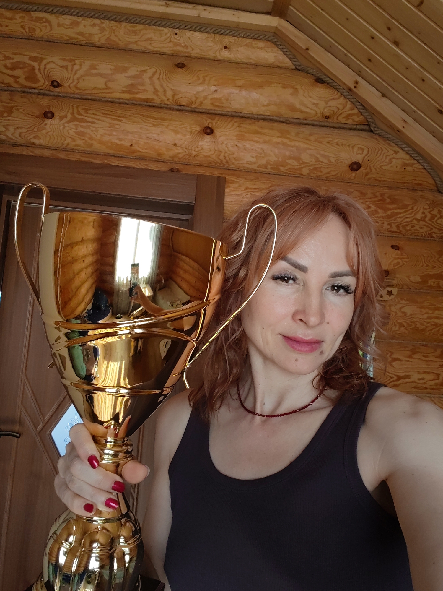 A woman holds a large gold trophy in front of a wooden paneled wall. The woman has light red hair, light skin, and is wearing a dark tank top and a red beaded necklace. She is holding the trophy with her right hand, which has red nail polish. The trophy is highly reflective, showing a distorted reflection of the surrounding environment. The wooden wall behind the woman appears to be made of logs. The location is likely in a building with wooden construction. The city is Старое Михайловское, Russia.